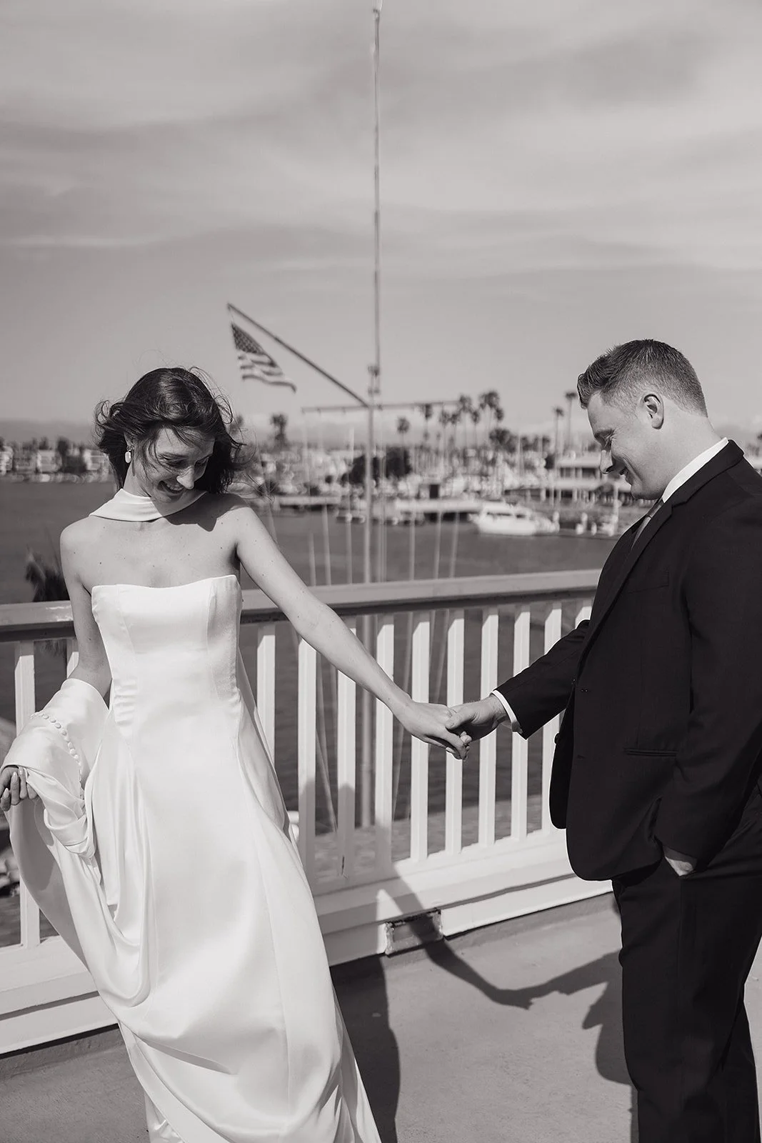 A bride and groom holding hands on a dock with boats and an American flag in the background, black and white photo.