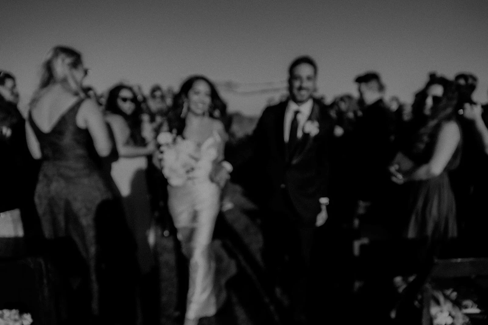 Black and white photo of a wedding party outdoors, featuring a bride and groom in the center surrounded by bridesmaids and groomsmen.