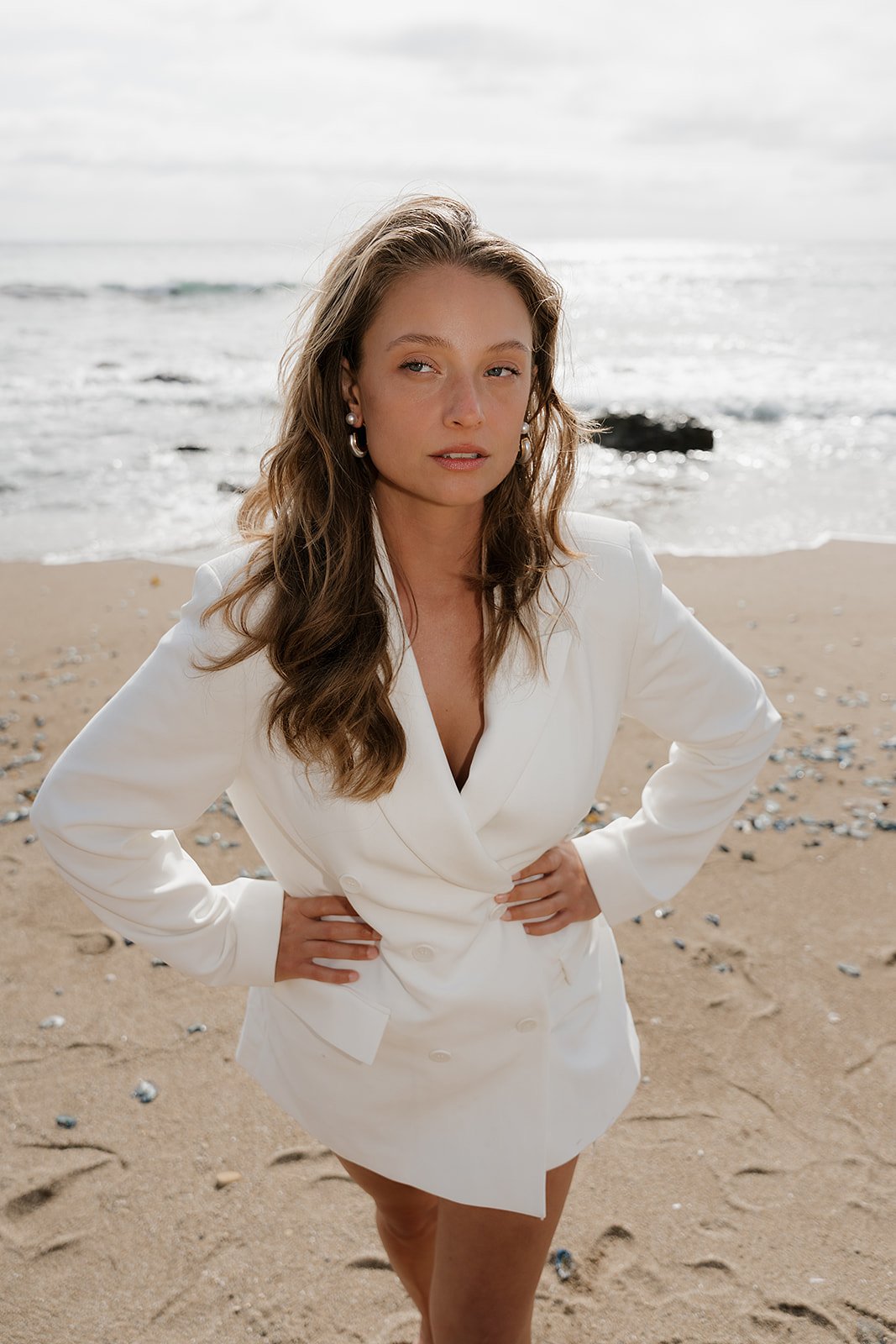Woman in a white blazer dress stands on a sandy beach with arms akimbo, facing the camera, with the ocean and cloudy sky in the background.