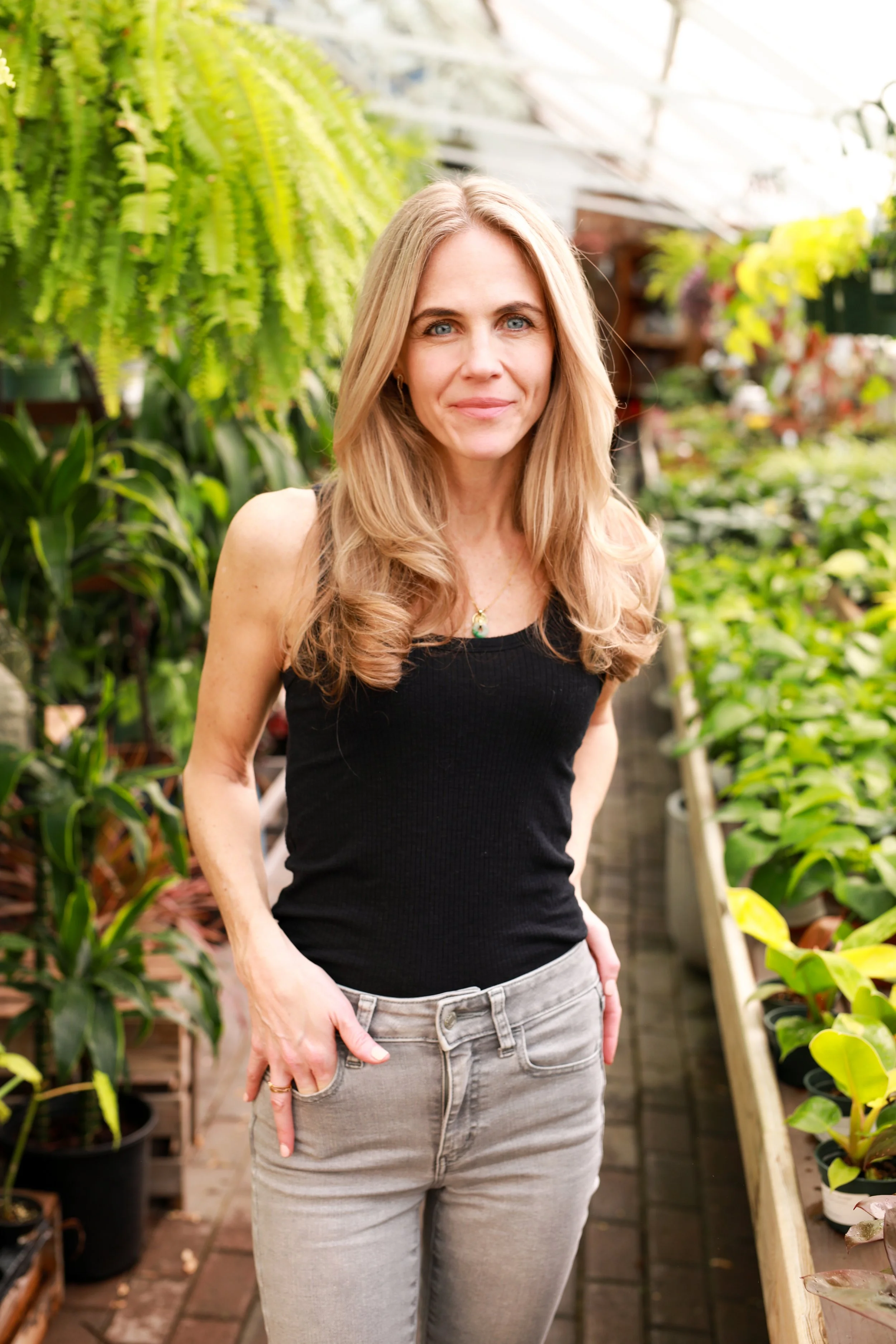 A woman with long blonde hair wearing a black sleeveless top and light gray pants standing in a greenhouse surrounded by potted plants and lush greenery.