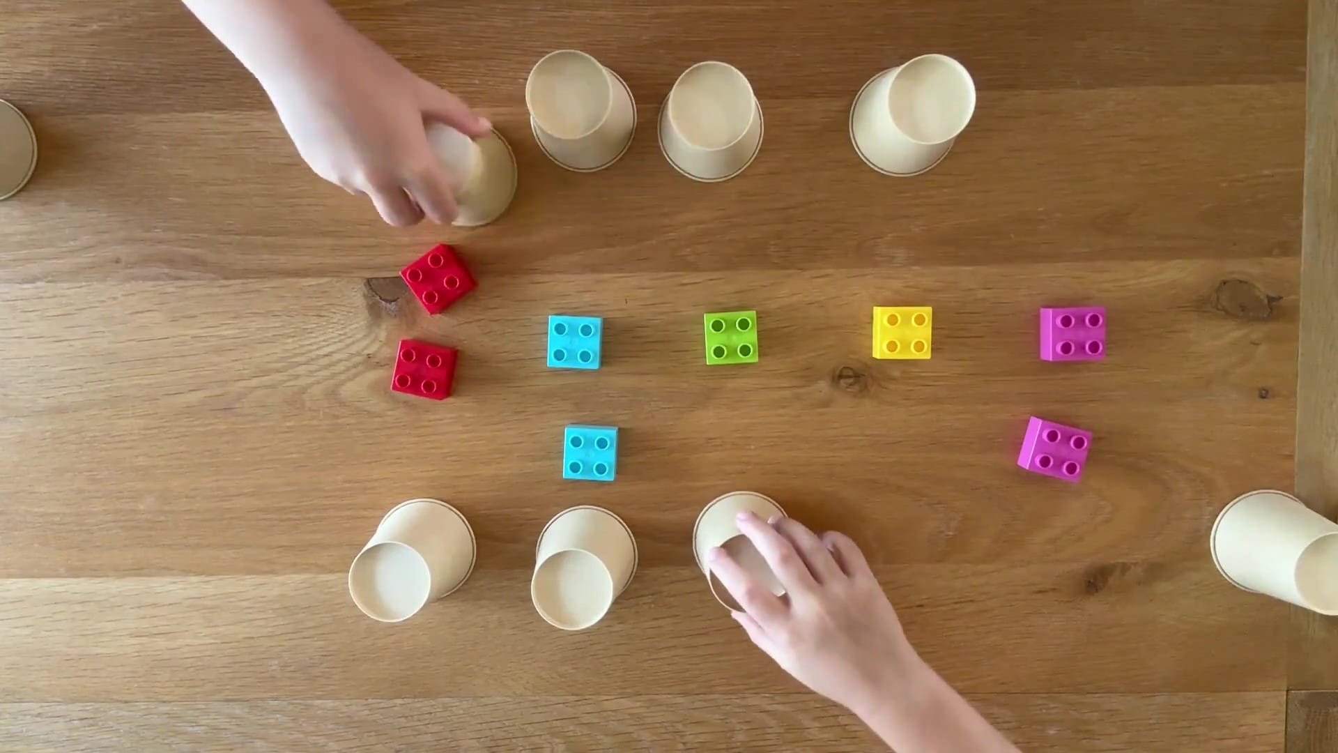 Children with hands on cups that have blocks under and moving them to the coloured duplo blocks in front of them to match them.