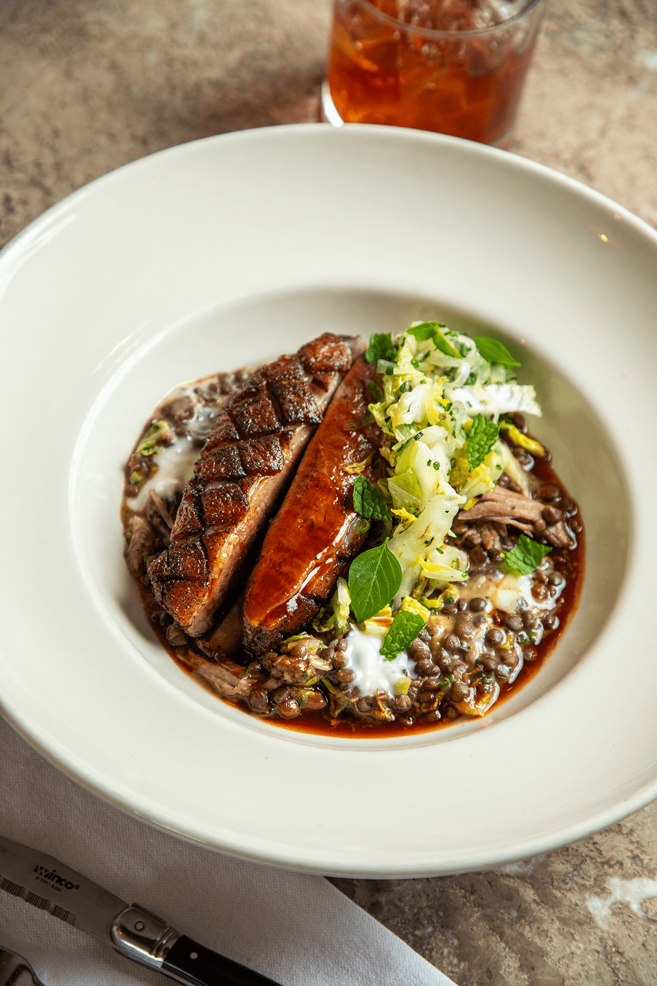 A white bowl containing a gourmet dish with sliced roasted meat, a glazed sausages, and a bed of lentils topped with chopped greens and herbs.