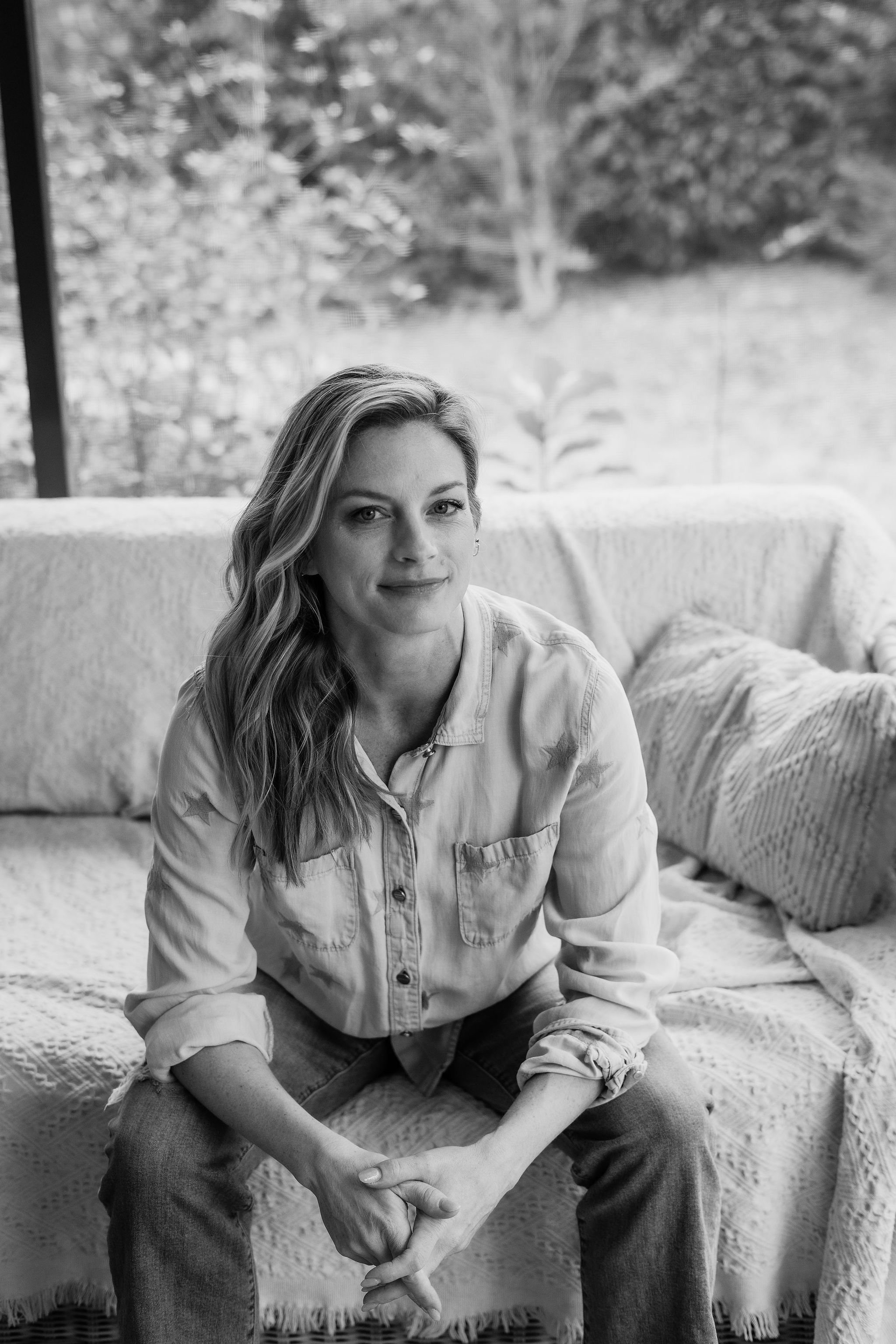 A woman with long wavy hair, wearing a collared shirt with rolled-up sleeves and ripped jeans, sitting on a bed with a textured blanket and pillows. She is indoors, in front of a large window with trees and foliage visible outside.
