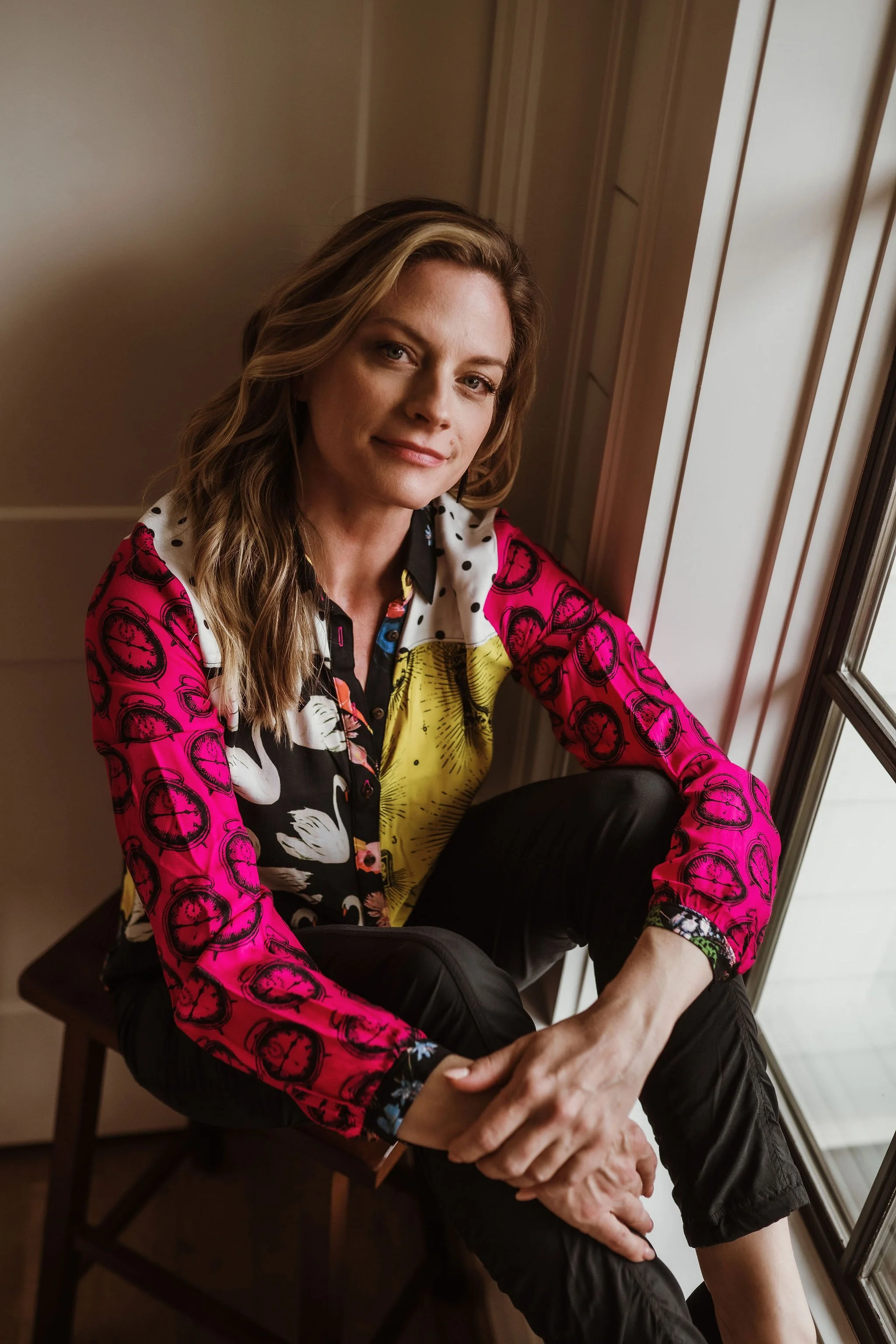 A woman with wavy brown hair and light complexion sitting by a window with natural light, wearing a colorful patterned shirt and black pants, looking at the camera with a gentle smile.