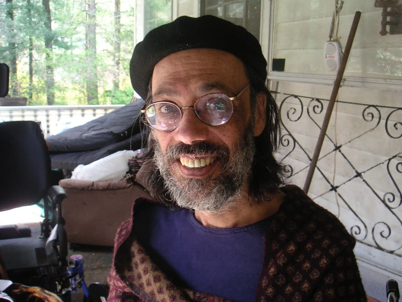 Smiling man with glasses and beard, wearing a black beret and a patterned jacket, outdoors on a porch with trees in the background.