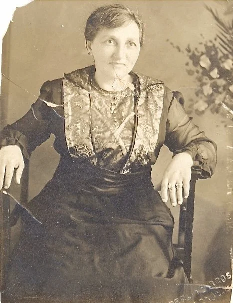 A black-and-white portrait of a woman sitting in a chair, wearing a dress with decorative fabric on the front, with a floral background on the right side.