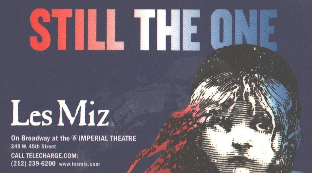 Poster advertising Les Miz on Broadway at the Imperial Theatre, featuring an illustration of a young girl's face with long hair. The text reads "STILL THE ONE" with "Les Miz" and theater details below.