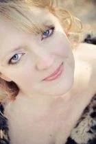 Close-up of a woman with fair skin, blonde hair, and blue eyes, smiling gently, with a blurred background.