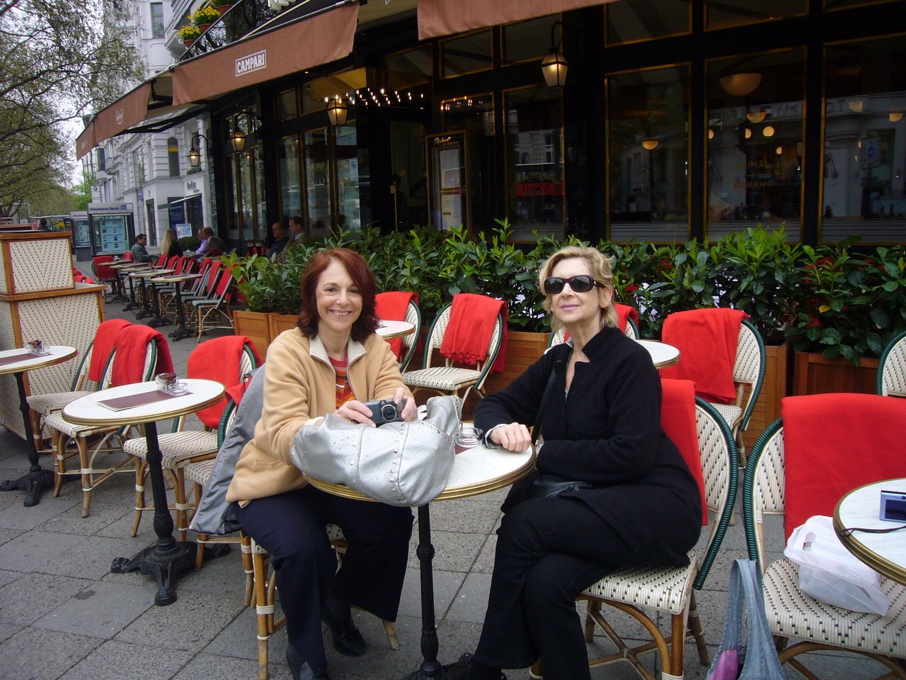 Two women sitting at an outdoor café table with red chairs and umbrellas, one holding a camera, on a city sidewalk.
