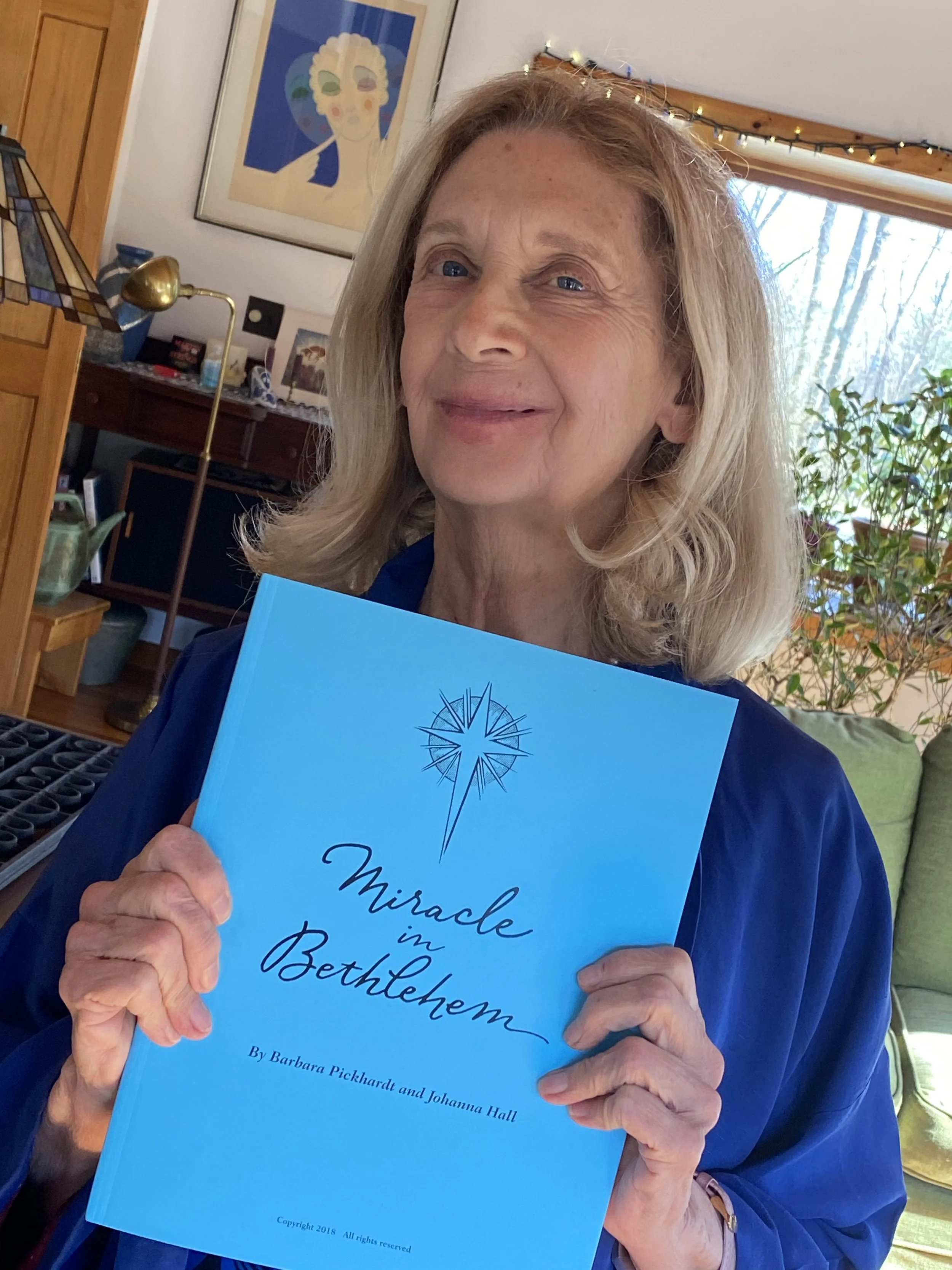 An elderly woman with blonde hair holding a blue book titled 'Miracle in Bethlehem' by Barbara Pickhardt and Johanna Hall, inside a cozy living room with artwork, a lamp, and plants in the background.