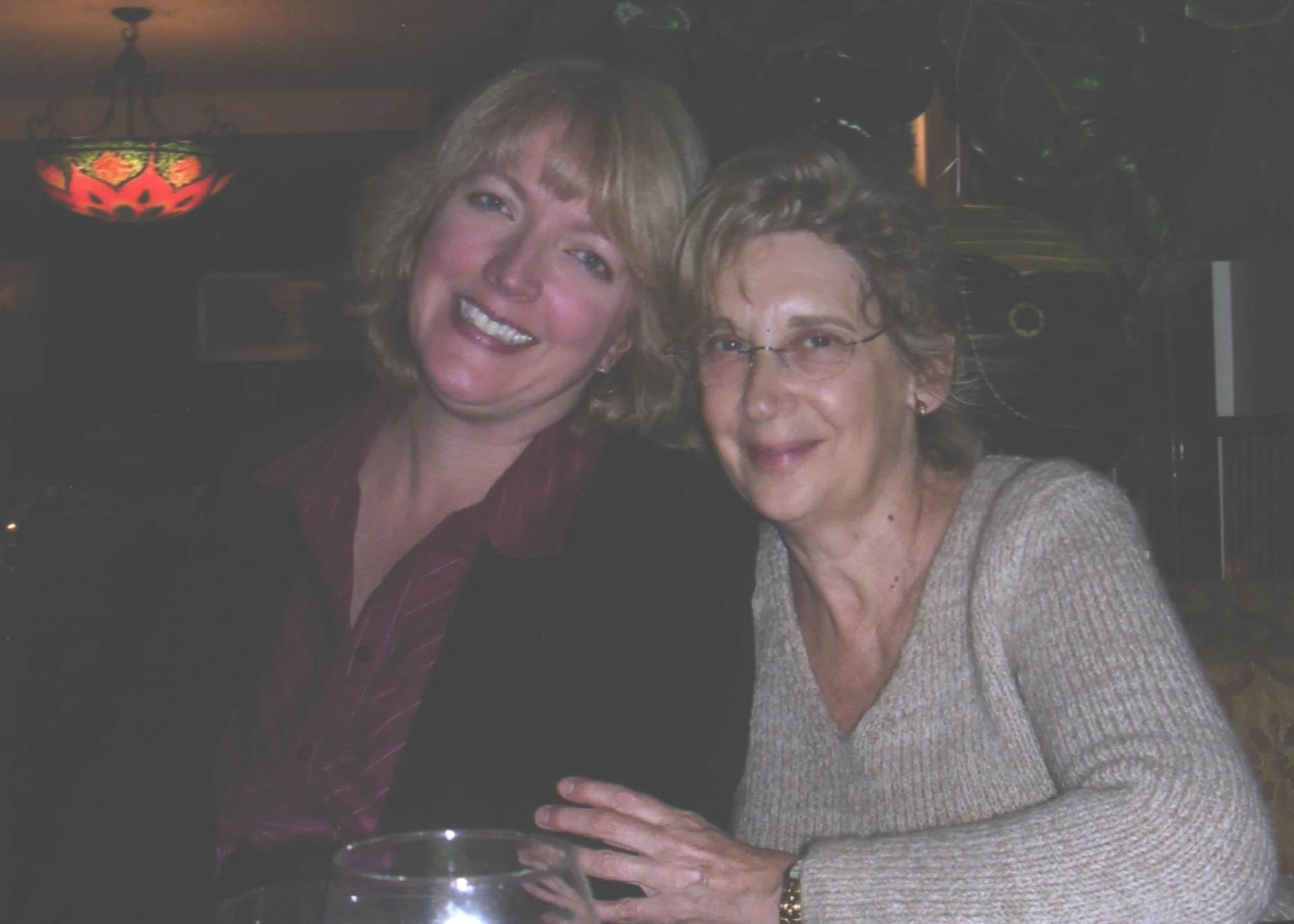 Two women smiling together at a social gathering, sitting close with their arms around each other, in a dimly lit room with colorful hanging lamps and plants in the background.
