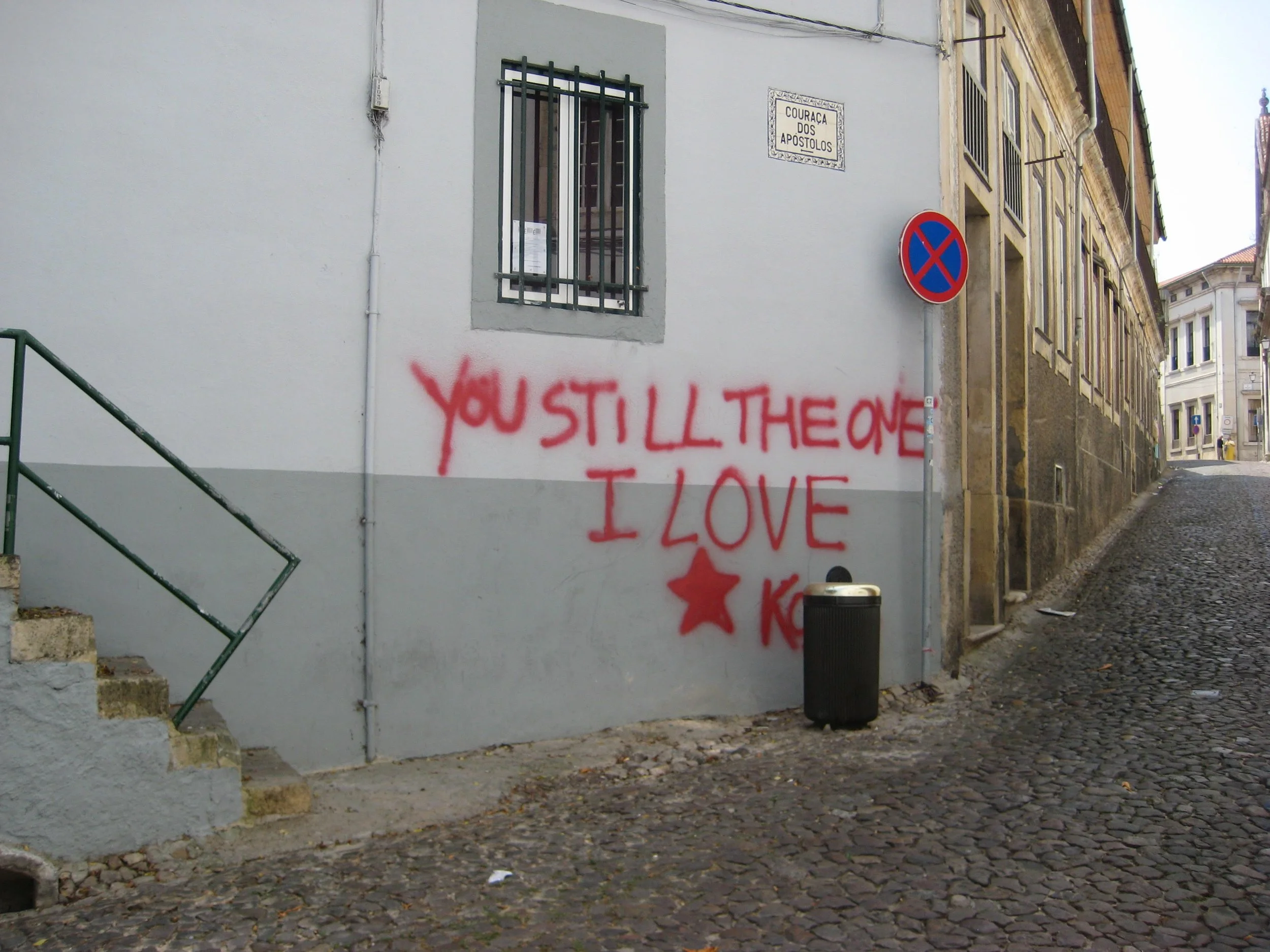 Graffiti on a white wall with the message 'You still the one I love' written in red spray paint, near a window with metal bars and a no-parking sign on a narrow cobblestone street.