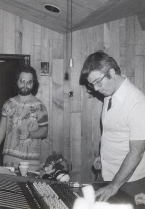 Two men in a recording studio, one is operating a mixing console while the other stands nearby, holding a cup. The room has wood-paneled walls and is equipped with recording equipment and microphones.