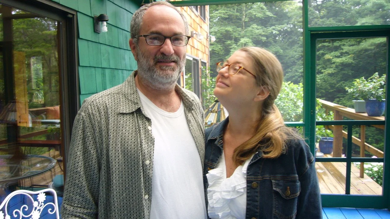 A man and woman standing outdoors on a porch with green walls and large windows, surrounded by trees and plants.