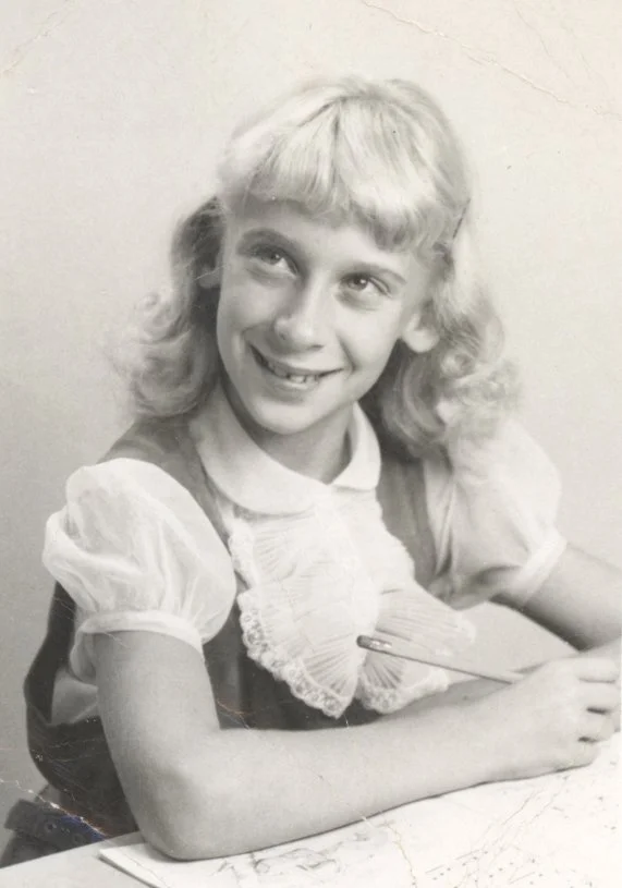 A smiling young girl with curly hair wearing a light-colored dress and a scalloped bib, sitting at a table with her arms resting on it, in a black-and-white photo.