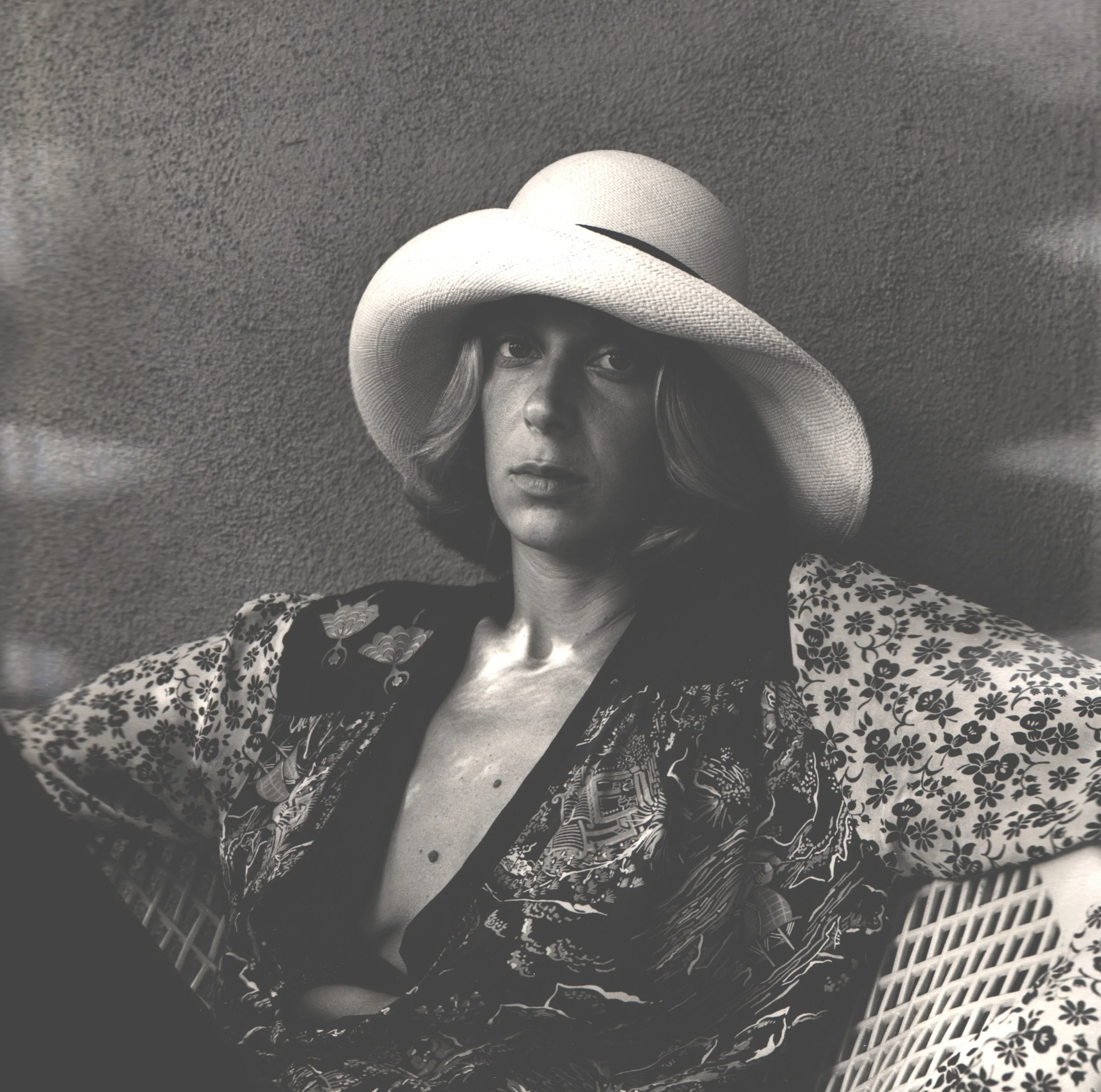 A woman wearing a large wide-brimmed hat and a patterned robe sitting on a chair with a textured wall behind her