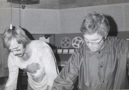 Two people working together at a desk in a recording studio with reels and equipment in the background.