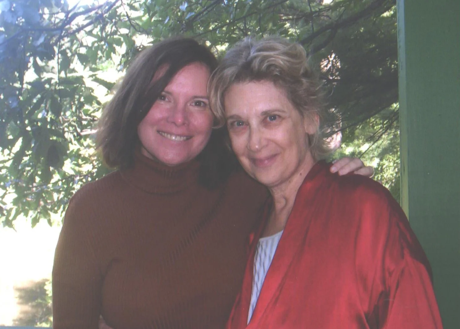Two women standing close together outdoors, smiling at the camera with a leafy green background.