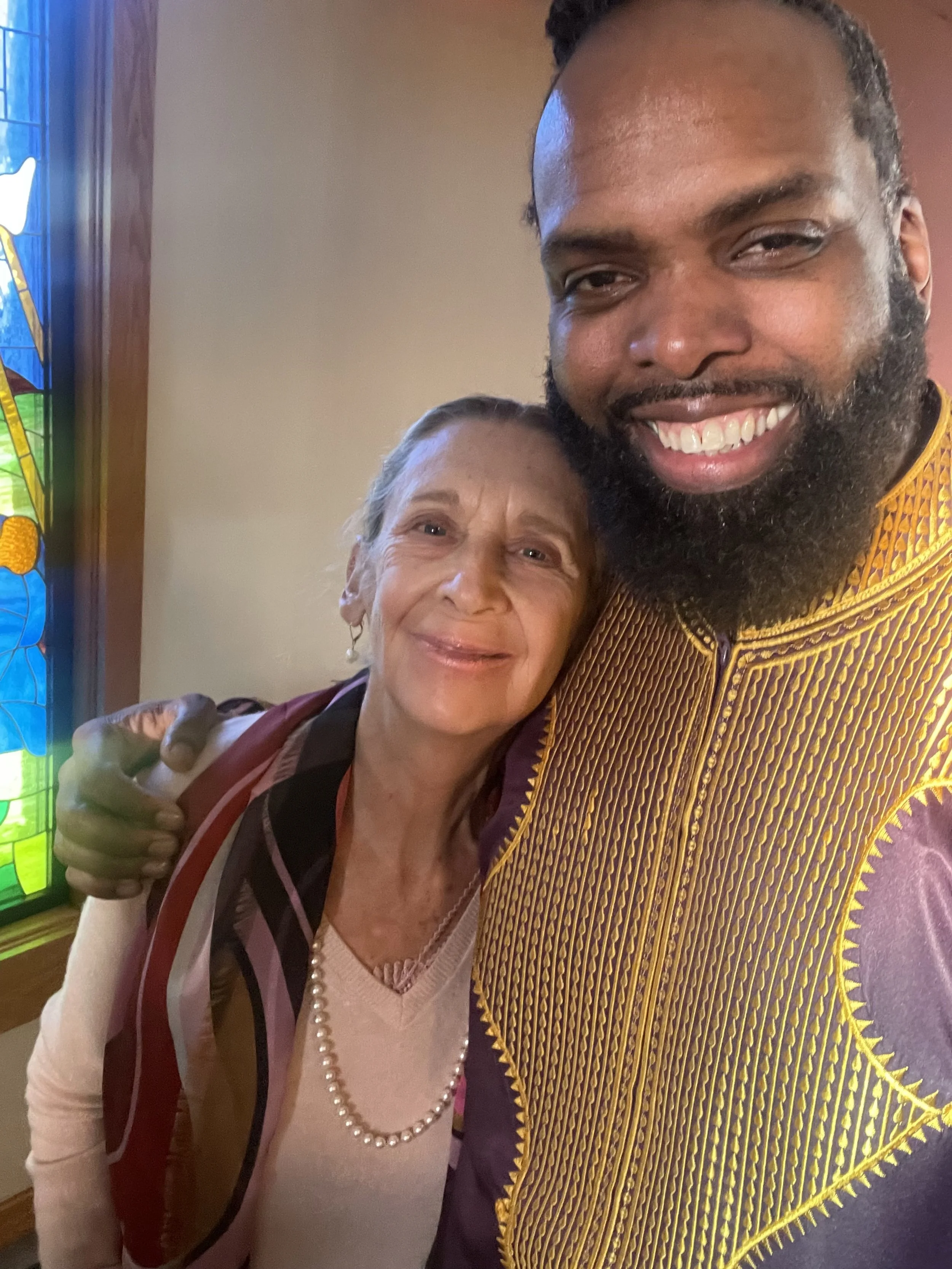 A young man and an elderly woman taking a selfie inside a church near a stained glass window. The young man has a beard and is smiling, wearing a traditional African patterned shirt. The elderly woman is also smiling, wearing a pearl necklace, and a light-colored top with a multicolored scarf.
