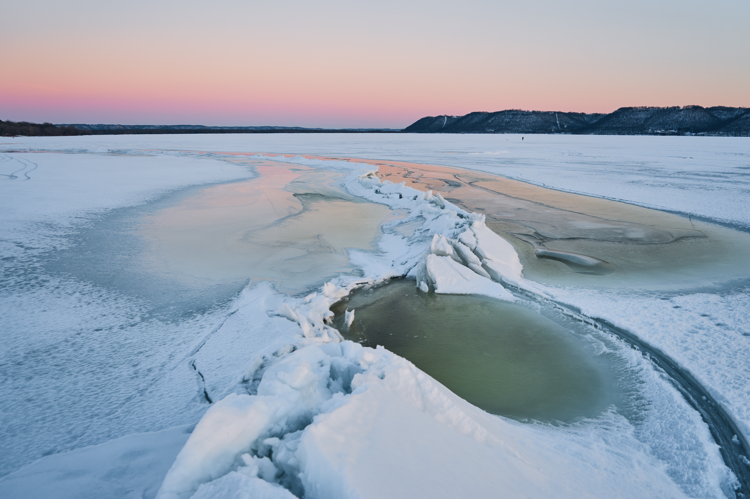 Frozen lake landscape with cracks and snow under a pink sunset sky.