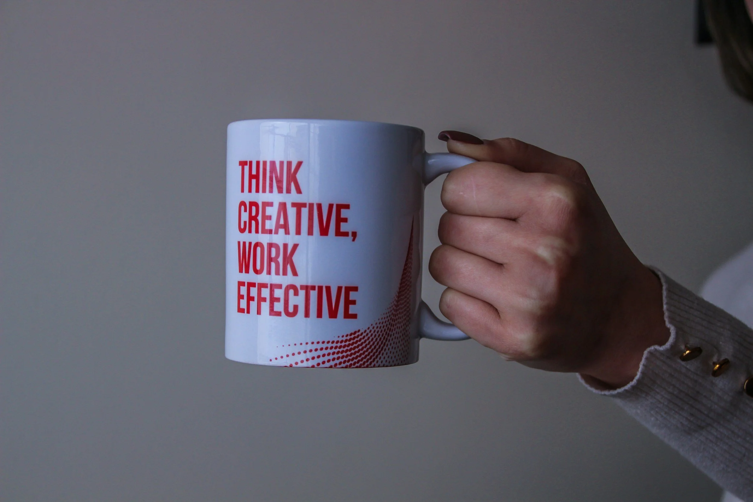 A person holding a white mug with red text that reads 'THINK CREATIVE, WORK EFFECTIVE' against a plain background.