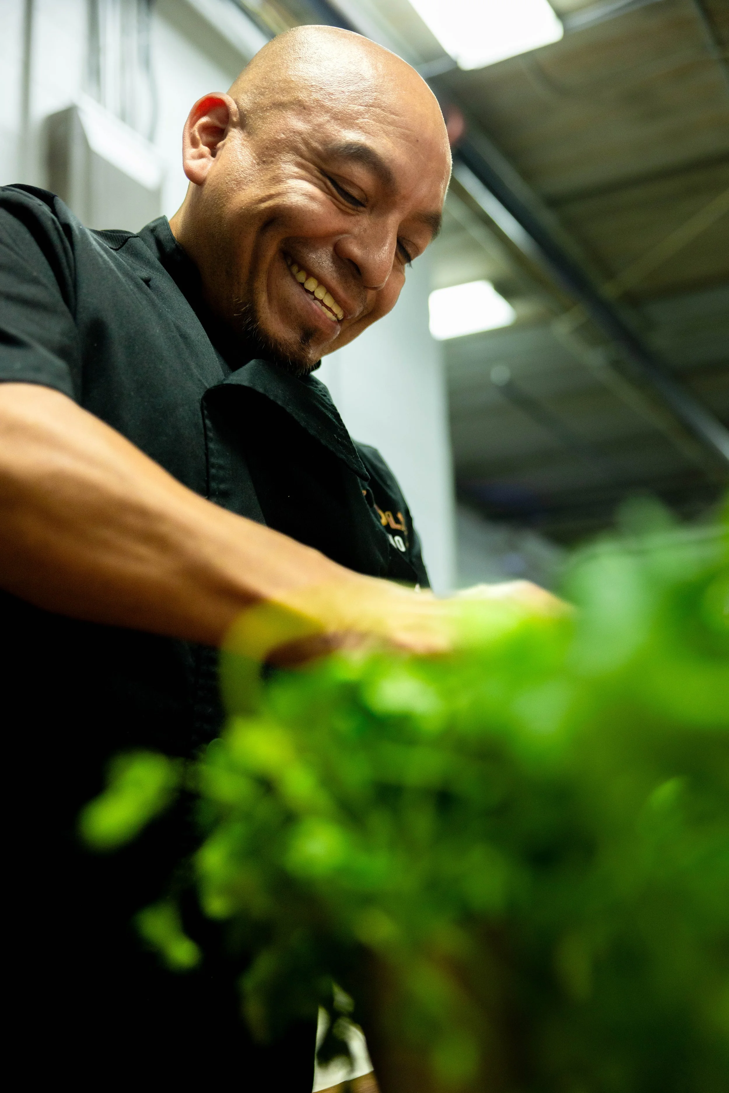 A man smiling while preparing food in a kitchen, wearing a black chef's coat.
