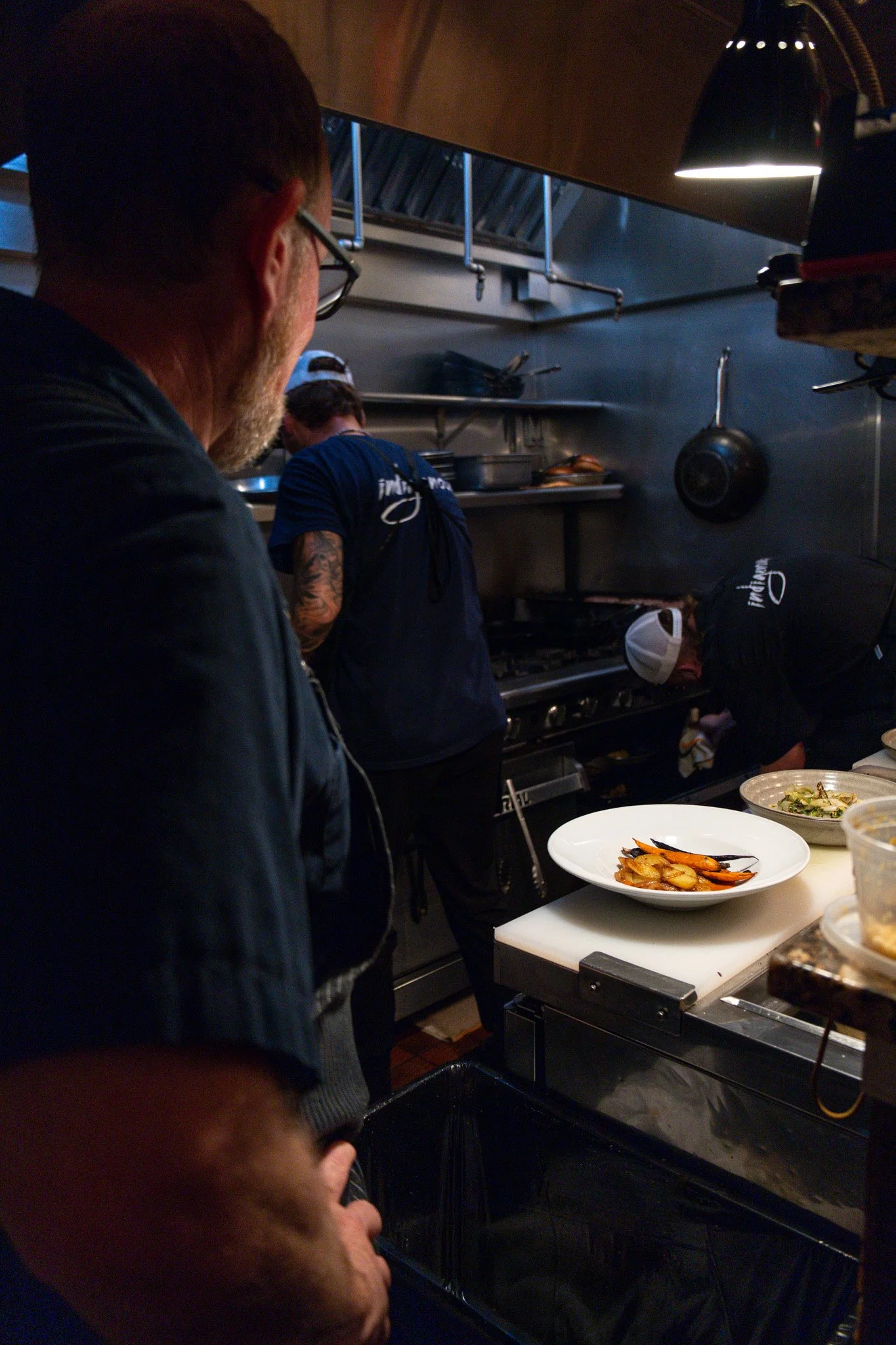 People working in a restaurant kitchen preparing food with plates of roasted vegetables on the counter.