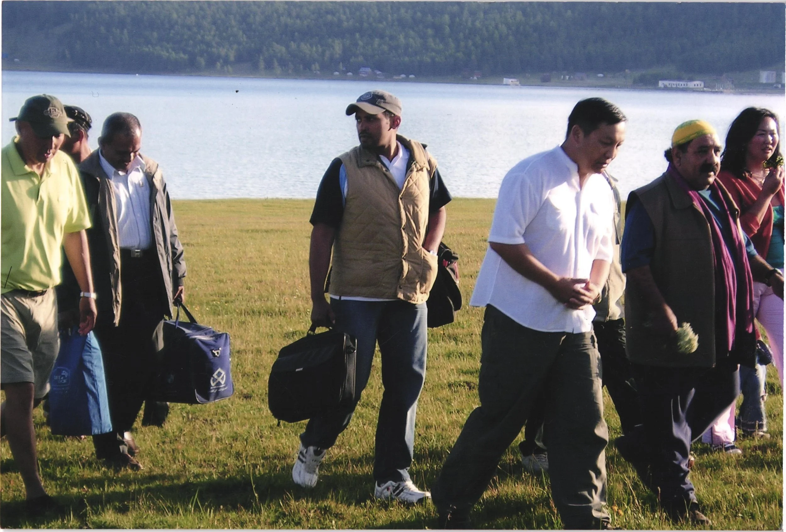 Group of diverse people walking outdoors near a body of water with a grassy field in the foreground and hills in the background.