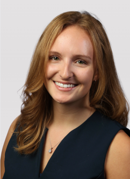A young woman with blonde hair and smiling and posing with one hand on her hip, wearing a sleeveless black top and black pants, standing against a plain light gray background.