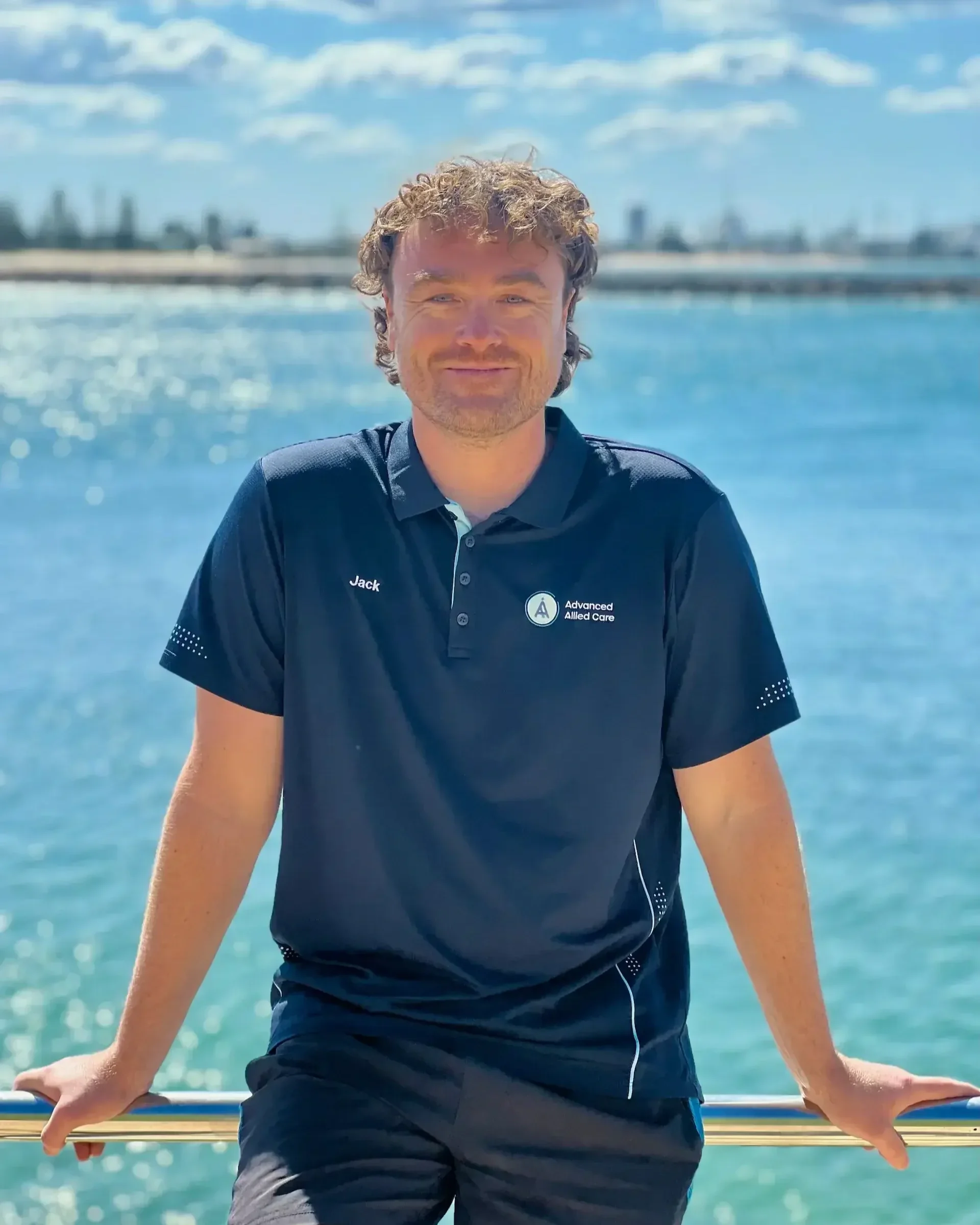 A man with curly hair smiling while sitting on a rail against a backdrop of the Newcastle harbour, blue sky, and distant city skyline.