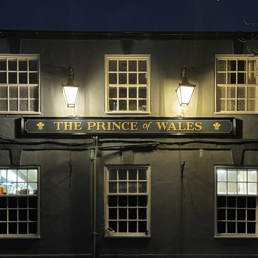 The exterior of a pub called 'The Prince of Wales' at night with lit lantern-style lights and several windows on a dark building facade.