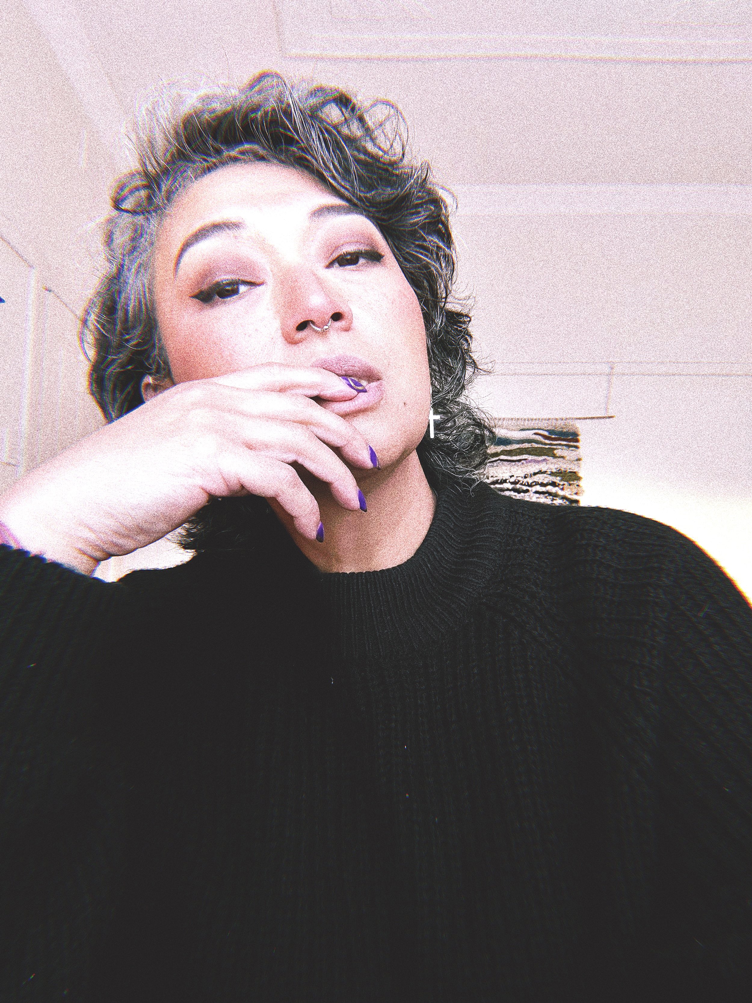 A woman with curly, dark hair, wearing a black sweater, with purple nail polish, resting her chin on her hand, in a room with a light-colored ceiling and a patterned wall art in the background.