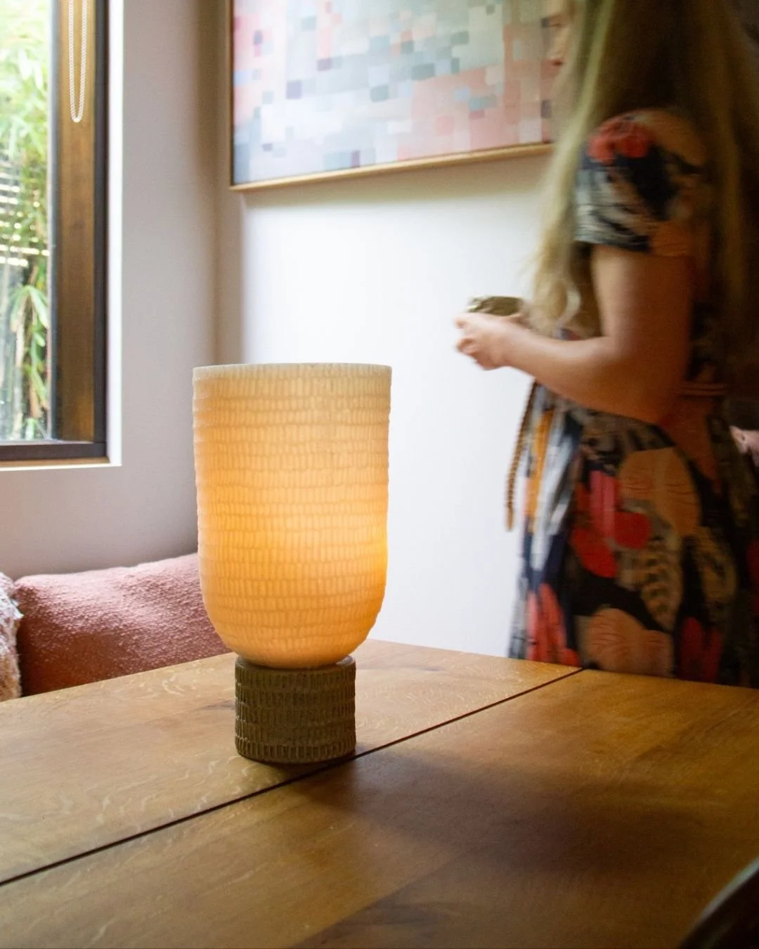 A beige textured lamp on a wooden table with a woman standing nearby, holding a coffee mug, in a room with a window and colorful artwork on the wall.