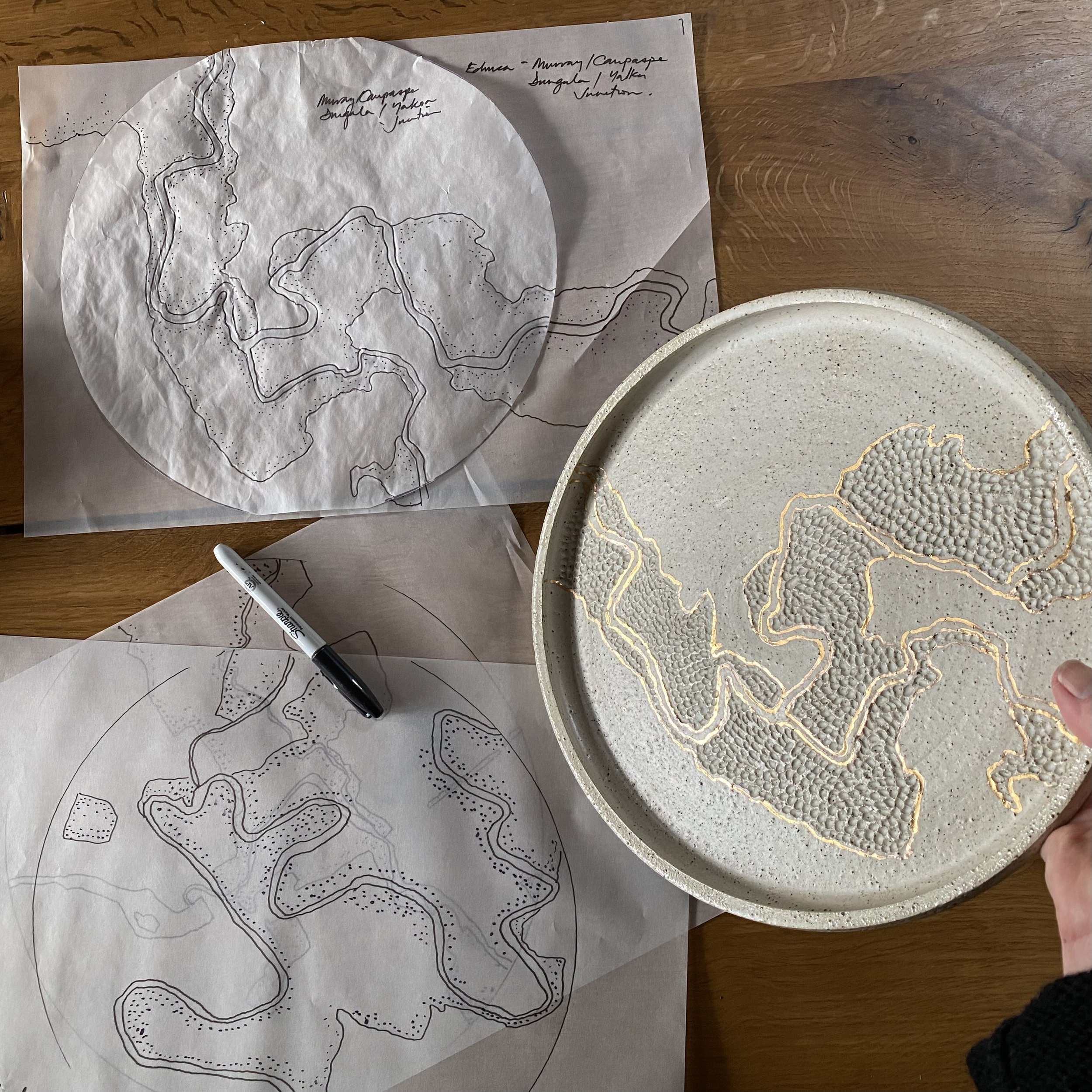 Hand-drawn topographical map on paper showing contour lines and land features, placed on a wooden surface, next to a decorative ceramic plate with gold accents, and a black marker.
