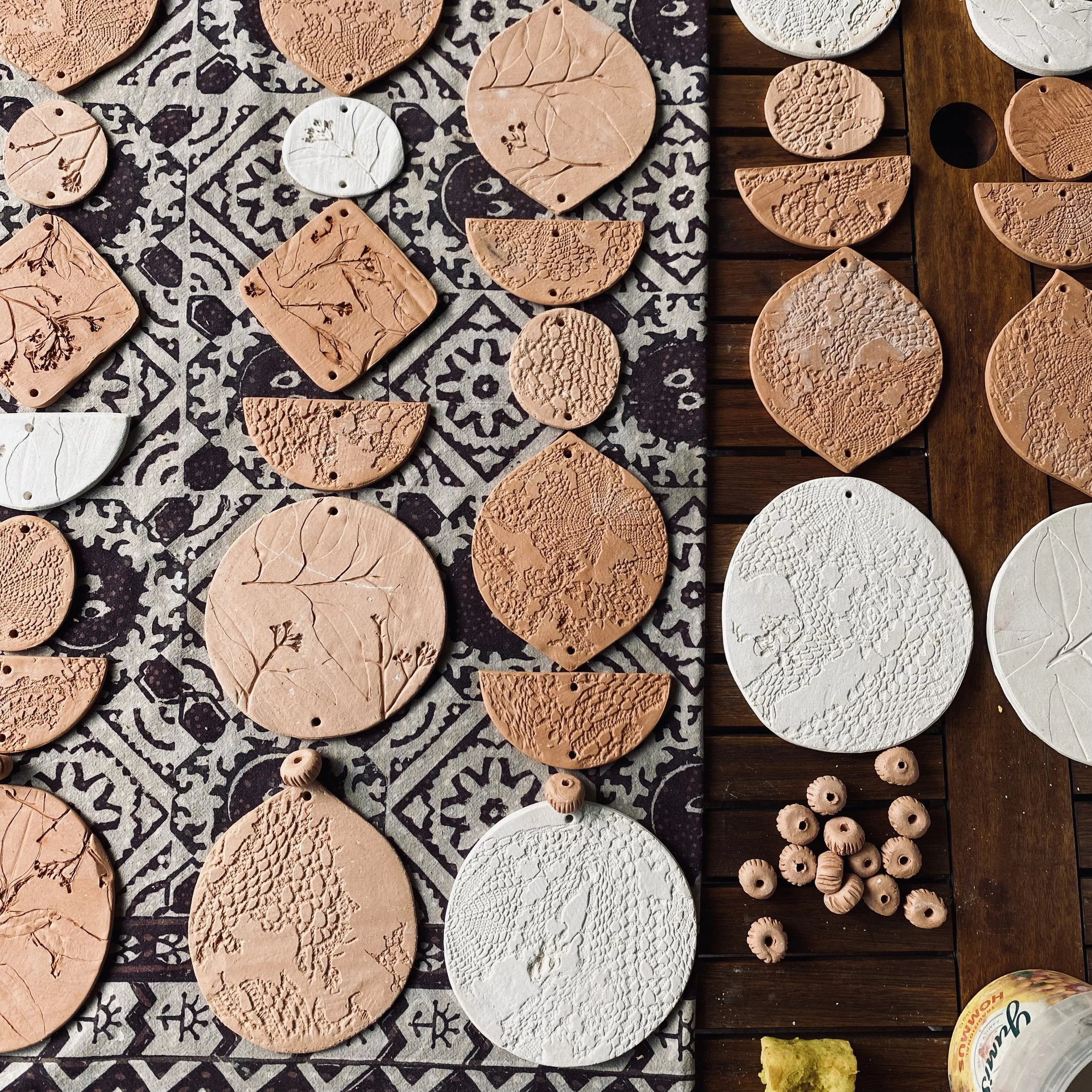 Clay or ceramic discs with engraved leaves and textured patterns, organized on a cloth and wooden surface, some with holes for hanging, and small clay beads nearby.