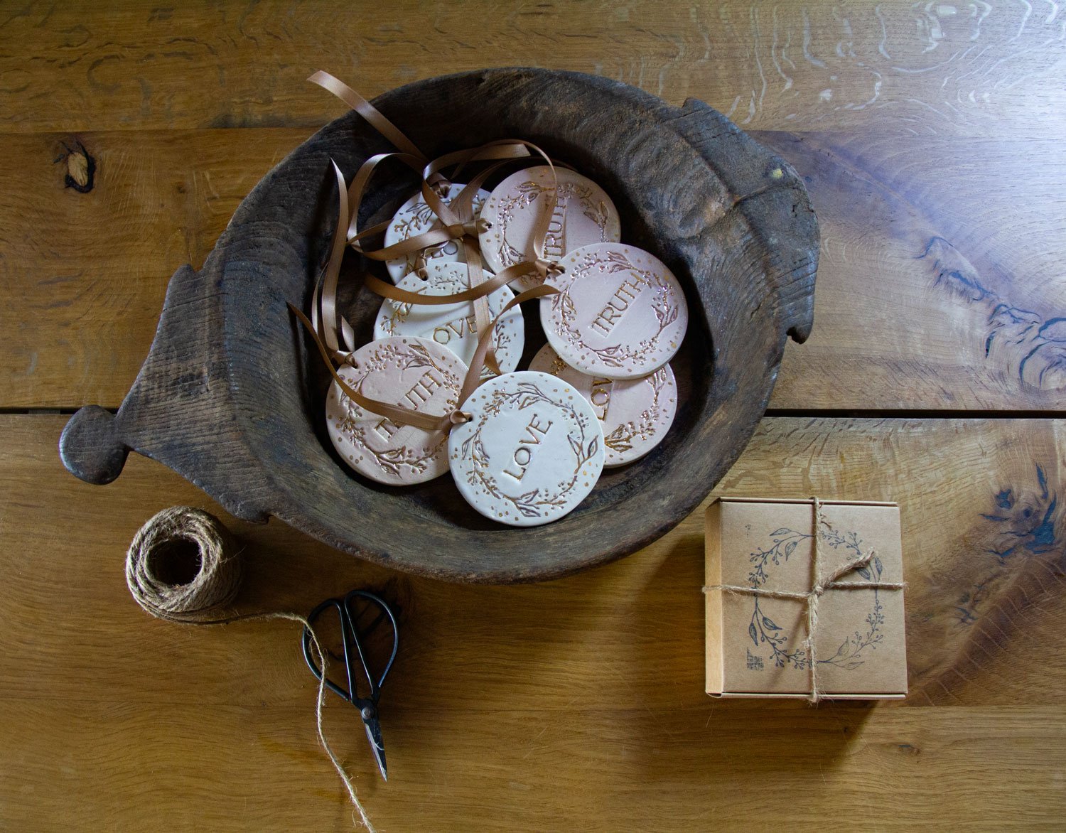 Round decorative ornaments with the word 'LOVE' on them, placed in a rustic wooden bowl. A small wrapped gift box and crafting supplies, including a spool of twine and scissors, are on a wooden surface.