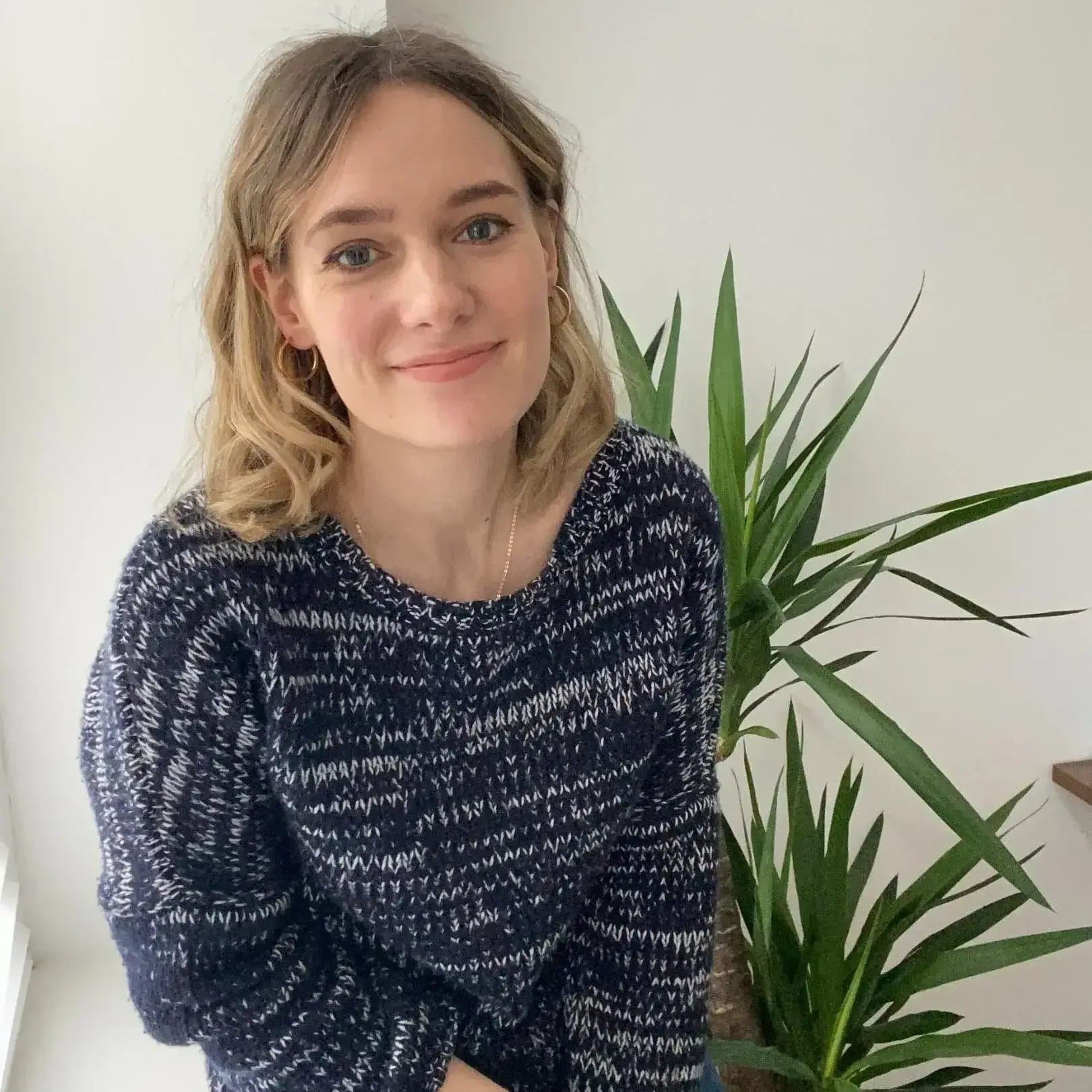 A woman with shoulder-length blonde hair and blue eyes, wearing a dark patterned sweater, smiling, standing in front of a green houseplant in a room with light-colored walls.