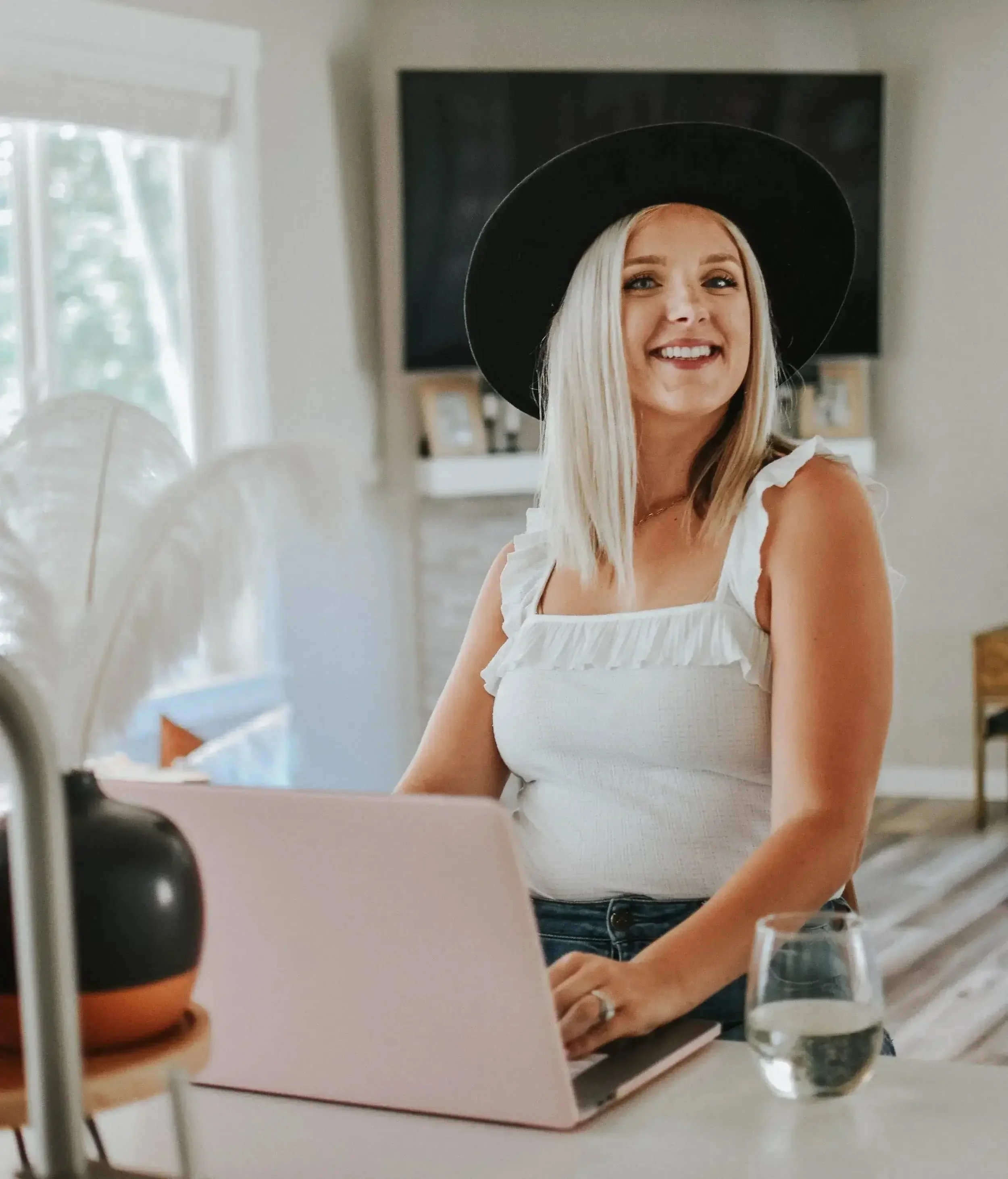 A woman smiling at a laptop in a bright room, wearing a white sleeveless top, a black wide-brimmed hat, and a ring on her finger, with a glass of water nearby.