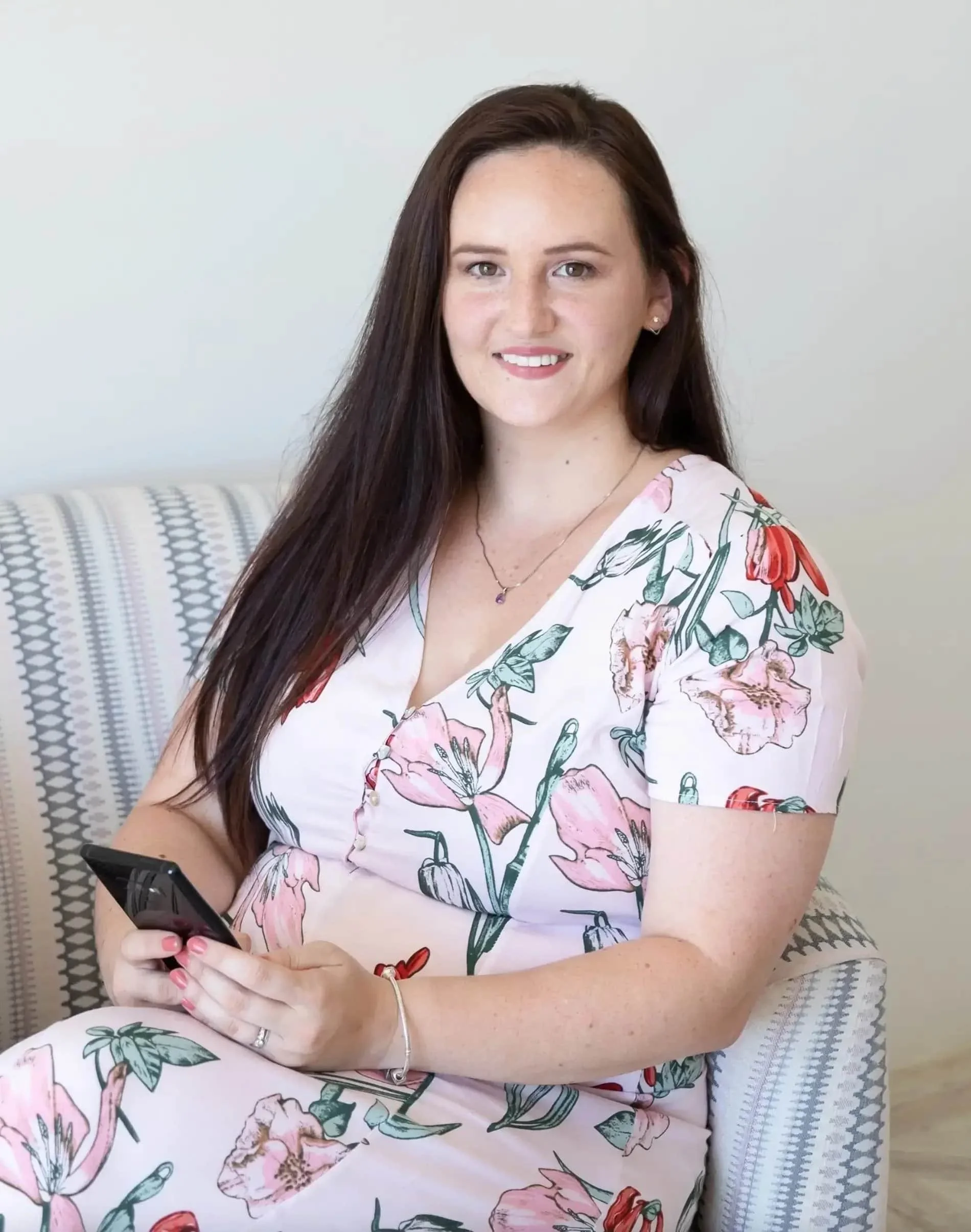 A woman with long dark hair sitting on a patterned sofa, wearing a floral dress and holding a smartphone.