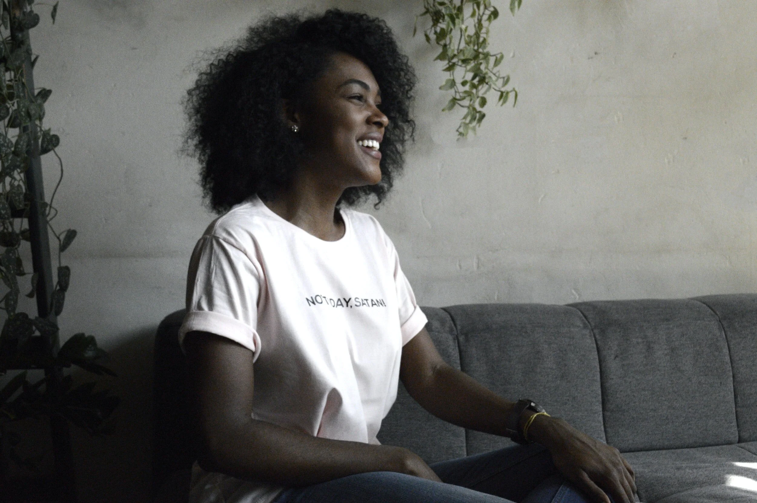 A young woman with curly black hair sitting on a gray couch, smiling and looking to her left, wearing a white T-shirt with the words 'NOT TODAY, SATAN!' printed on it, in a room with light-colored walls and some green plants.