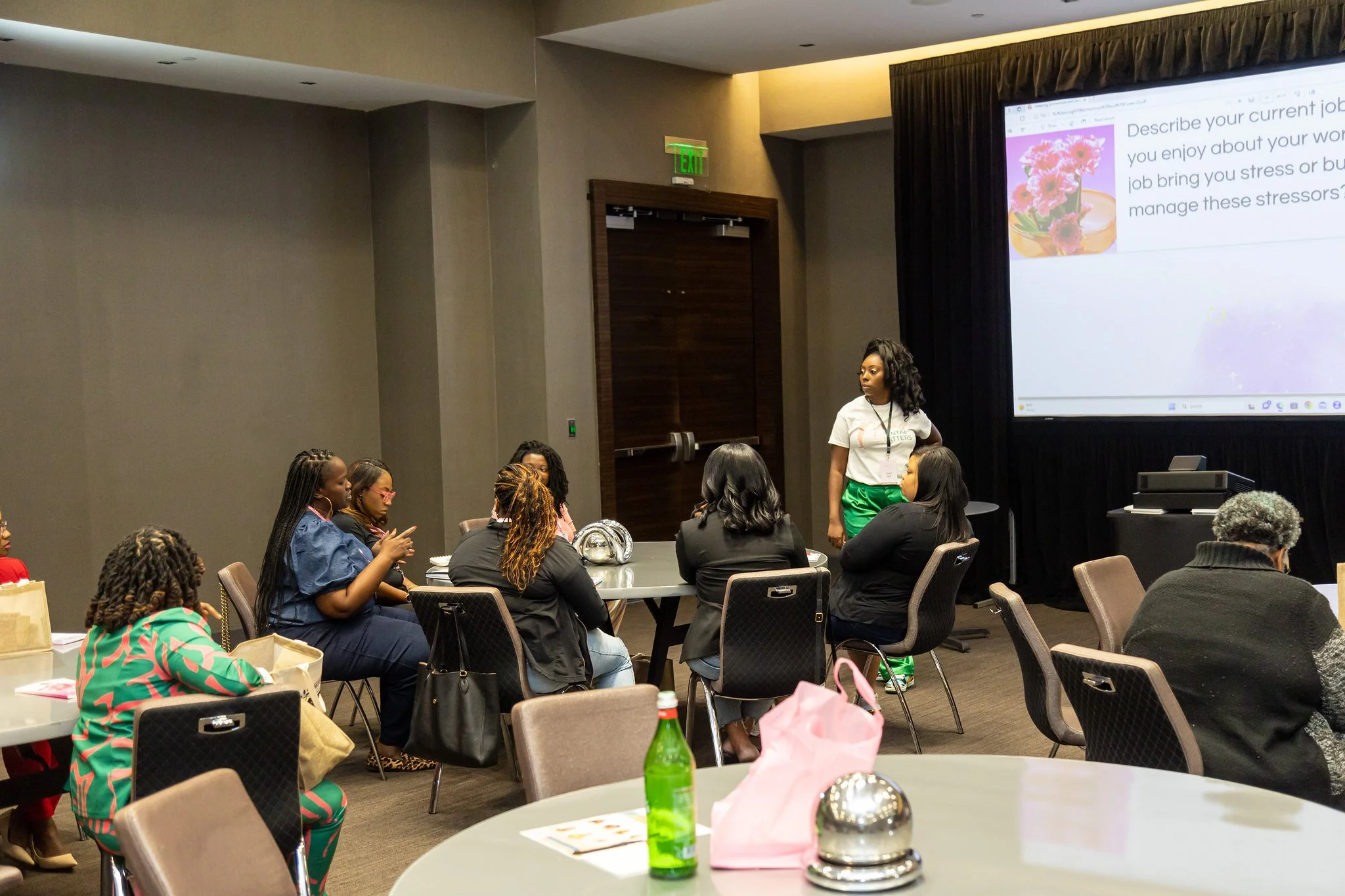 A woman standing next to a large screen in a conference room, giving a presentation to a seated audience of diverse women. The screen displays a slide about describing your current job and stress management. There are round tables with chairs, bottles, and bags in the foreground.