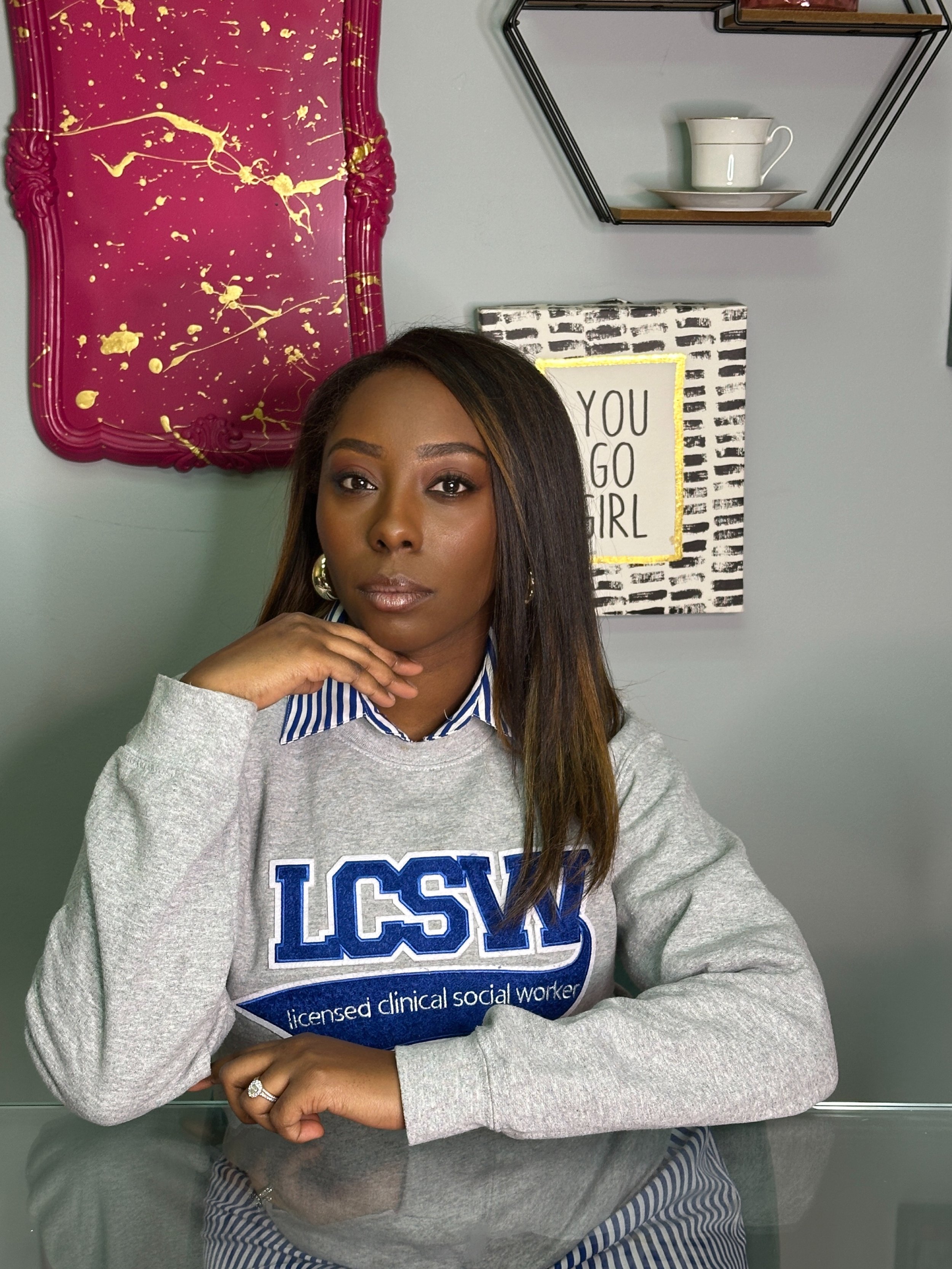 A woman sitting at a table in front of a light-colored wall with decorative decor, including a maroon and gold splatter tray, a photo with the text 'You Go Girl', and a hexagonal wall shelf with a teacup and saucer. The woman wears a gray sweatshirt with 'LCSW' and 'licensed clinical social worker' written on it, layered over a blue and white striped shirt, and has long dark hair.
