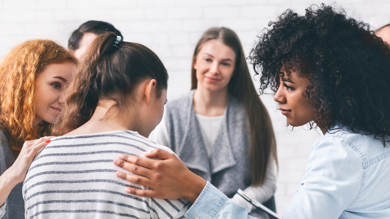 Group of women comforting and supporting a young girl who appears upset, in a room with white brick walls.