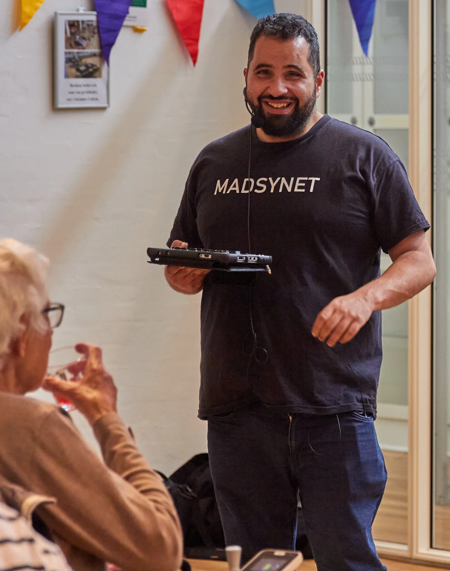 En mand med mørkt hår, skæg og et smil står med en mikrofon og holder en sort tablett i en indendørs lokal. Han bærer en sort T-shirt med teksten 'MADSNET' og har en headset mikrofon. I forgrunden ses en person med blond hår og briller, der spiser pizza.