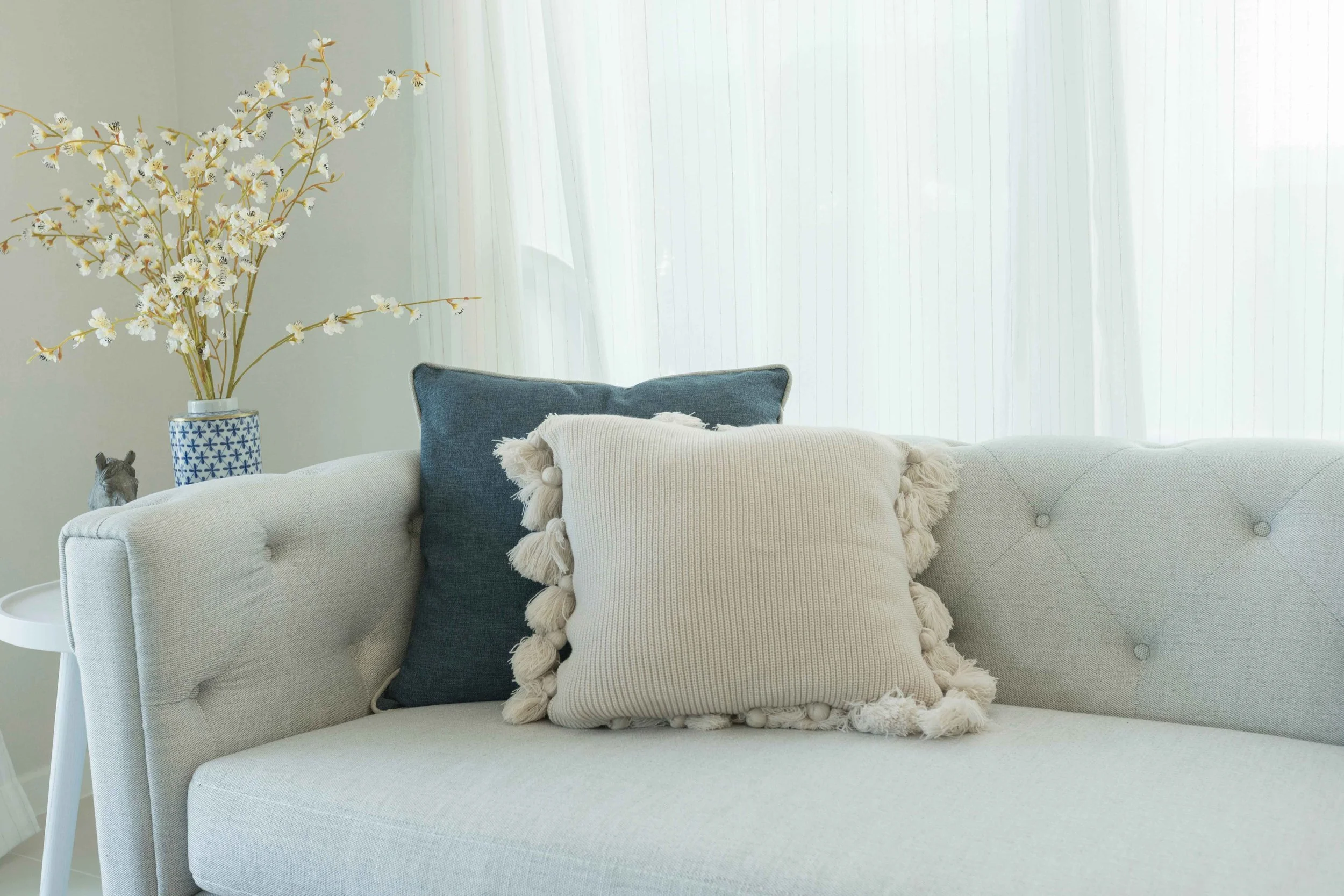 A neatly made bed with a blue upholstered headboard, white bedding, and decorative pillows including a large beige knitted pillow and two teal ones, beside a round wooden side table with a glass vase containing green foliage and a white woven table lamp.