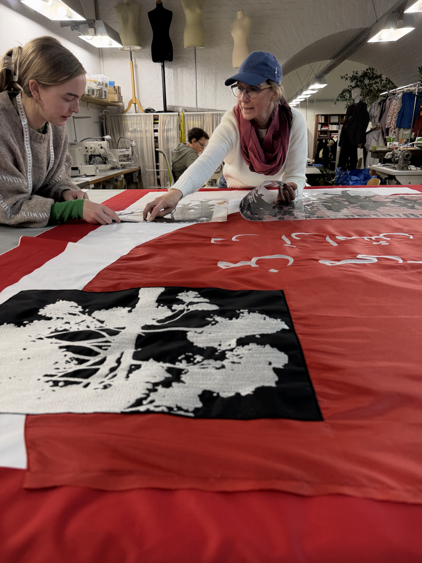 KÖR: Chefin Michaela und Lehrling Feli besprechen einen großen roten Banner, der auf dem Tisch ausgebreitet ist.