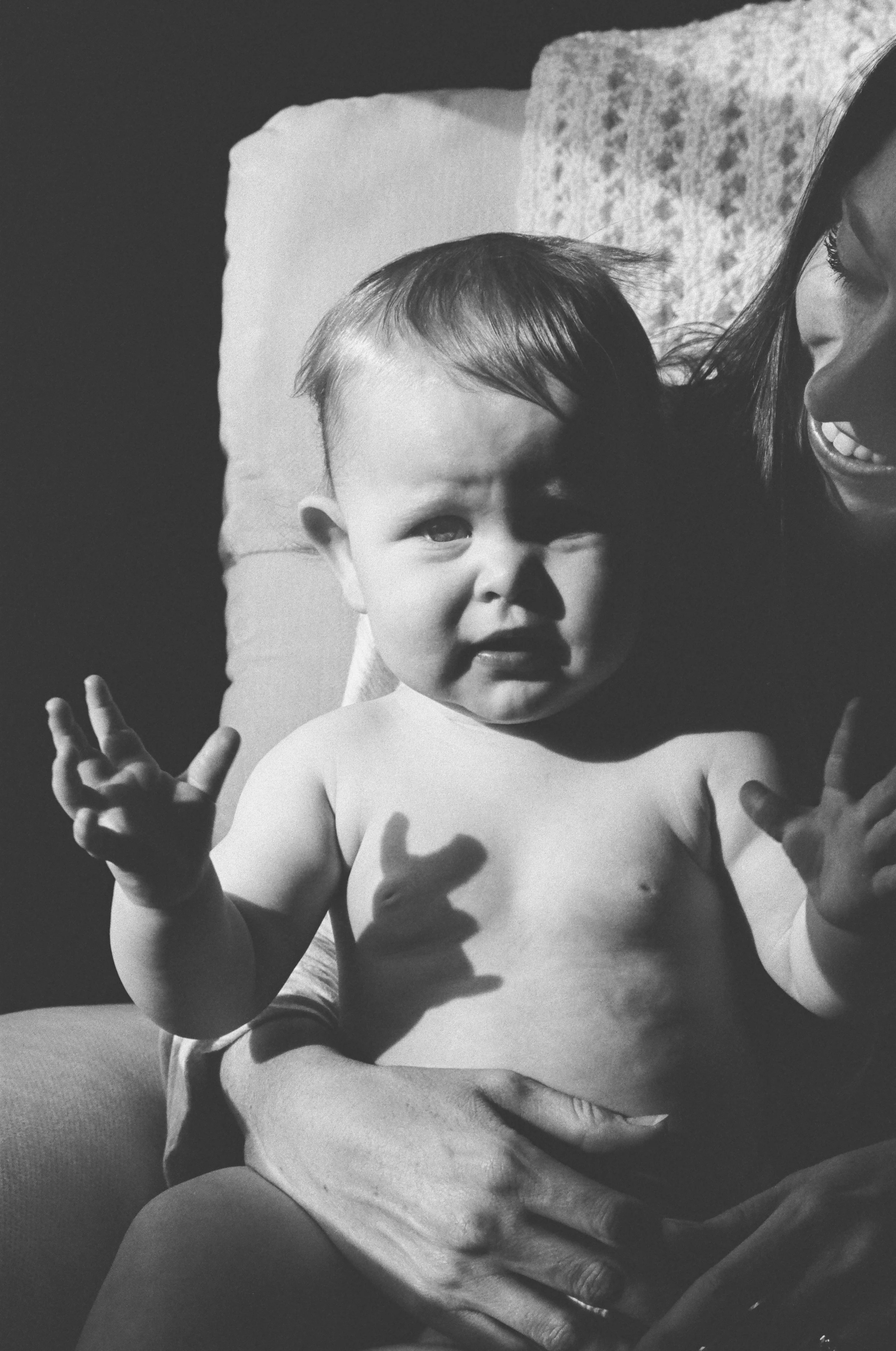 A black-and-white photo of a young child with a distressed expression lying on a person's lap. The child is shirtless, and the person is holding him with one hand on his chest. A woman smiling on 35mm film hp5.