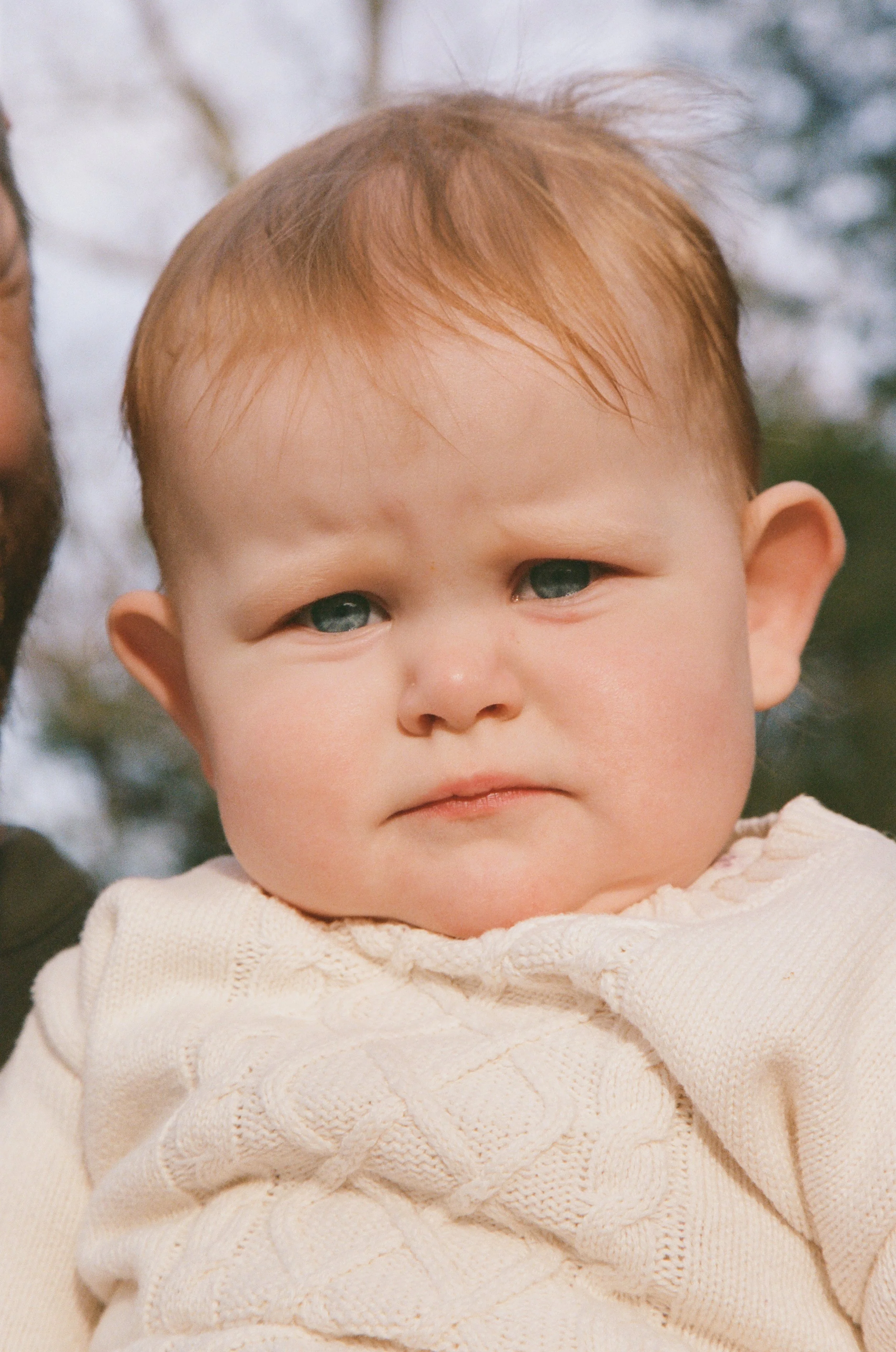 Close-up of a baby with fair skin, red hair, and blue eyes, wearing a cream-colored knitted sweater, outdoors with blurred trees in the background photographed on 35mm color analog film. 