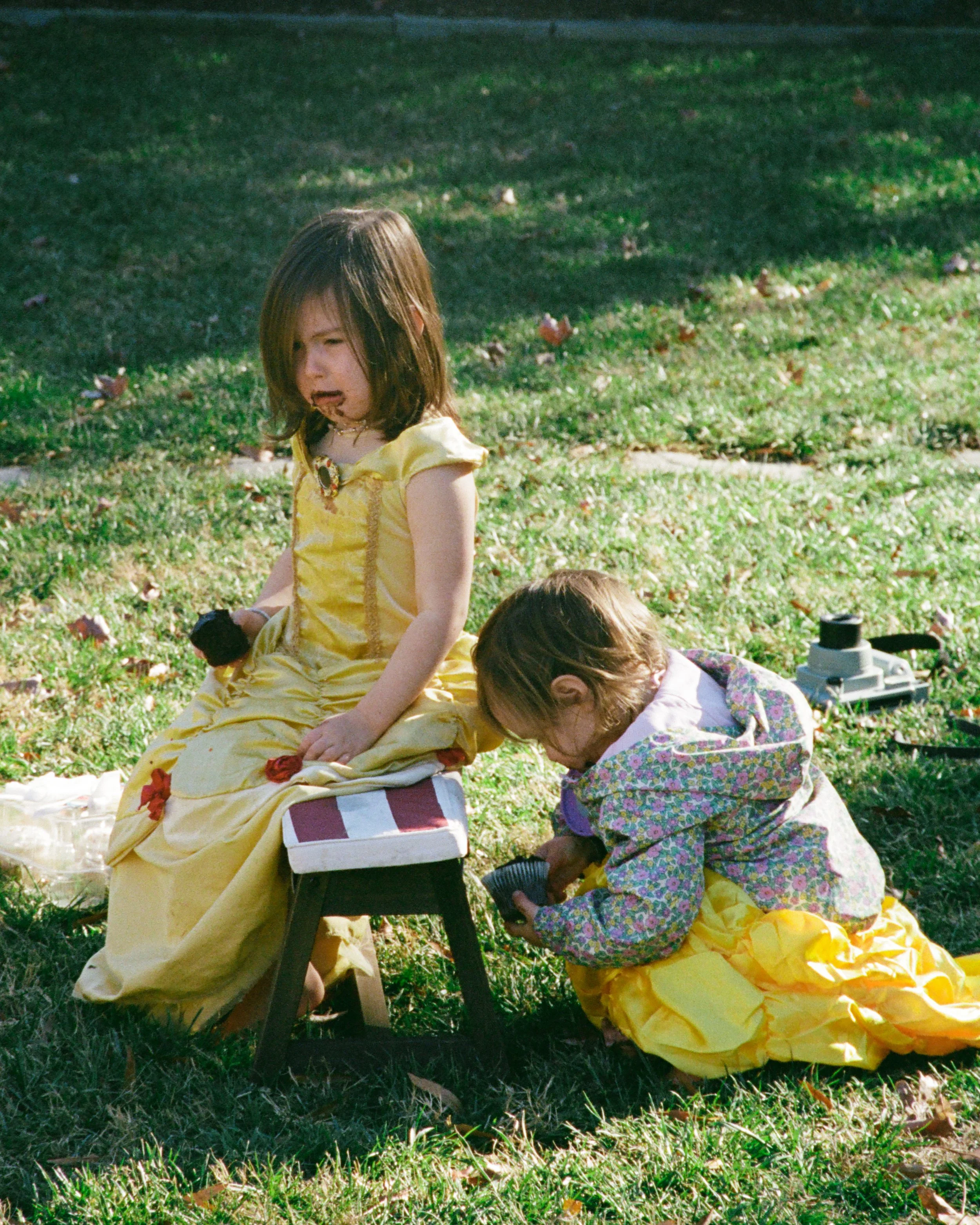 Two young girls in yellow princess dresses sitting on the grass outdoors, with one girl eating cupcakes and crying photographed on 35mm Kodak portra film 35mm in Richmond, Virginia 