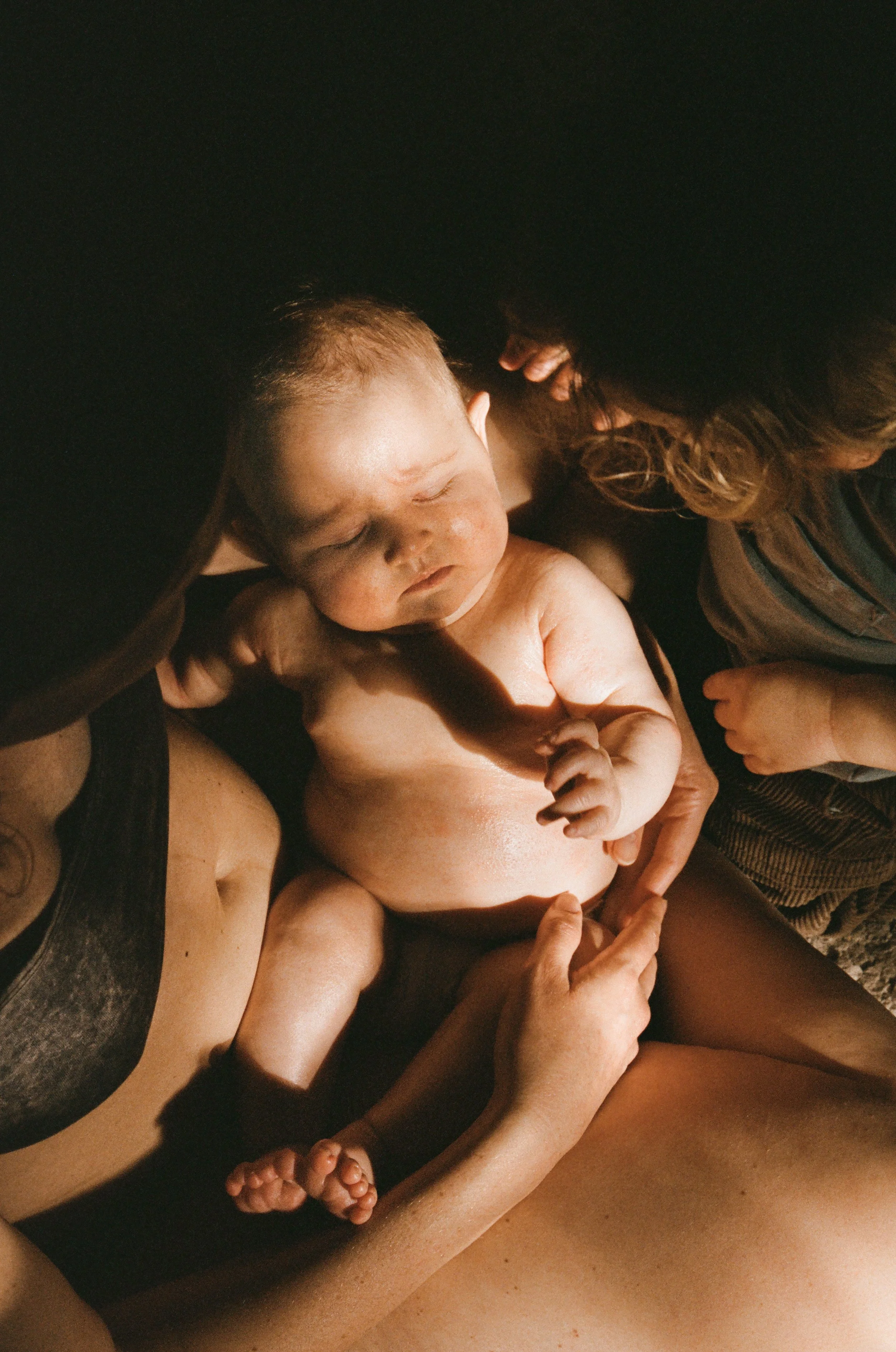A newborn baby resting peacefully on a woman's chest, with another child gently touching the baby's arm, in a warm and intimate moment captured on 35mm film for a postpartum newborn photoshoot. 